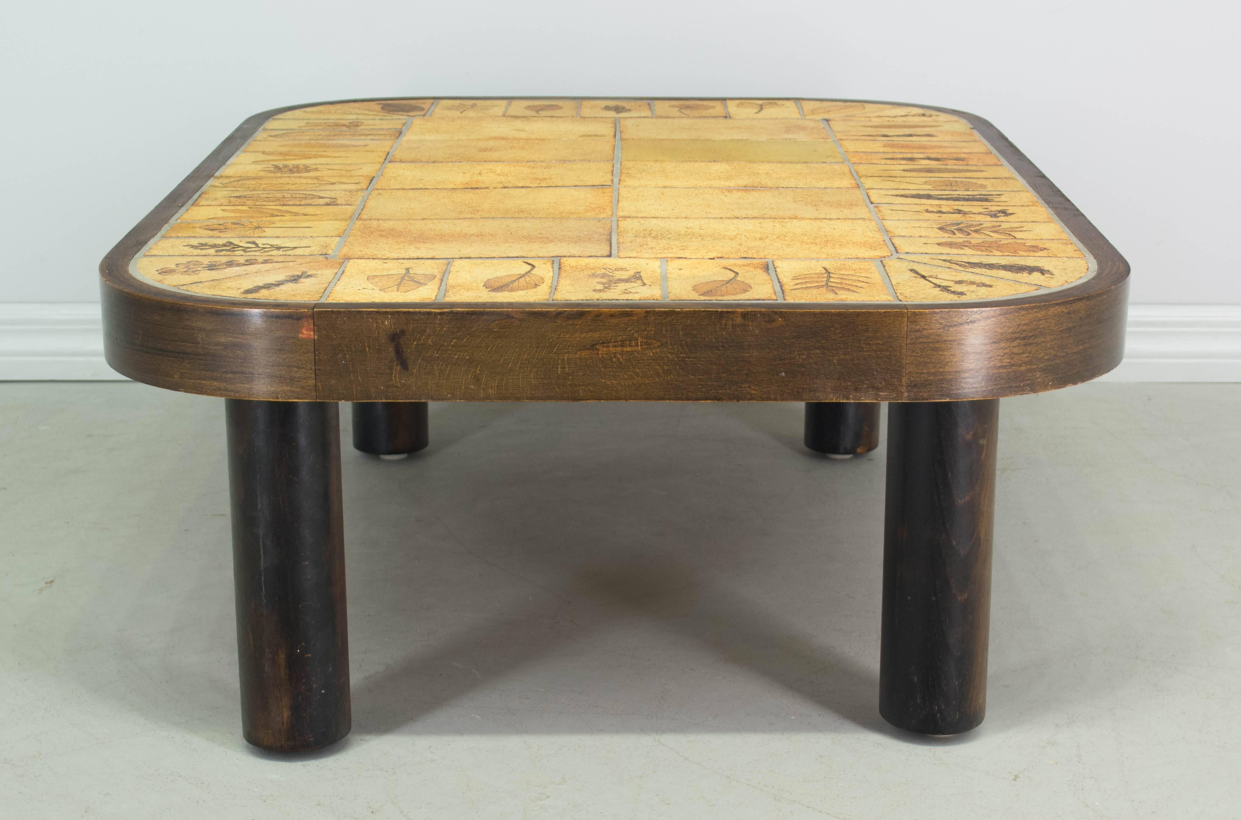 Midcentury French glazed ceramic tile top coffee table designed by Roger Capron. Fossil-like pressed leaves in the surface of the tiles that rim the perimeter. Rectangular wood base with curved corners and thick cylinder legs. Signed: R. Capron As