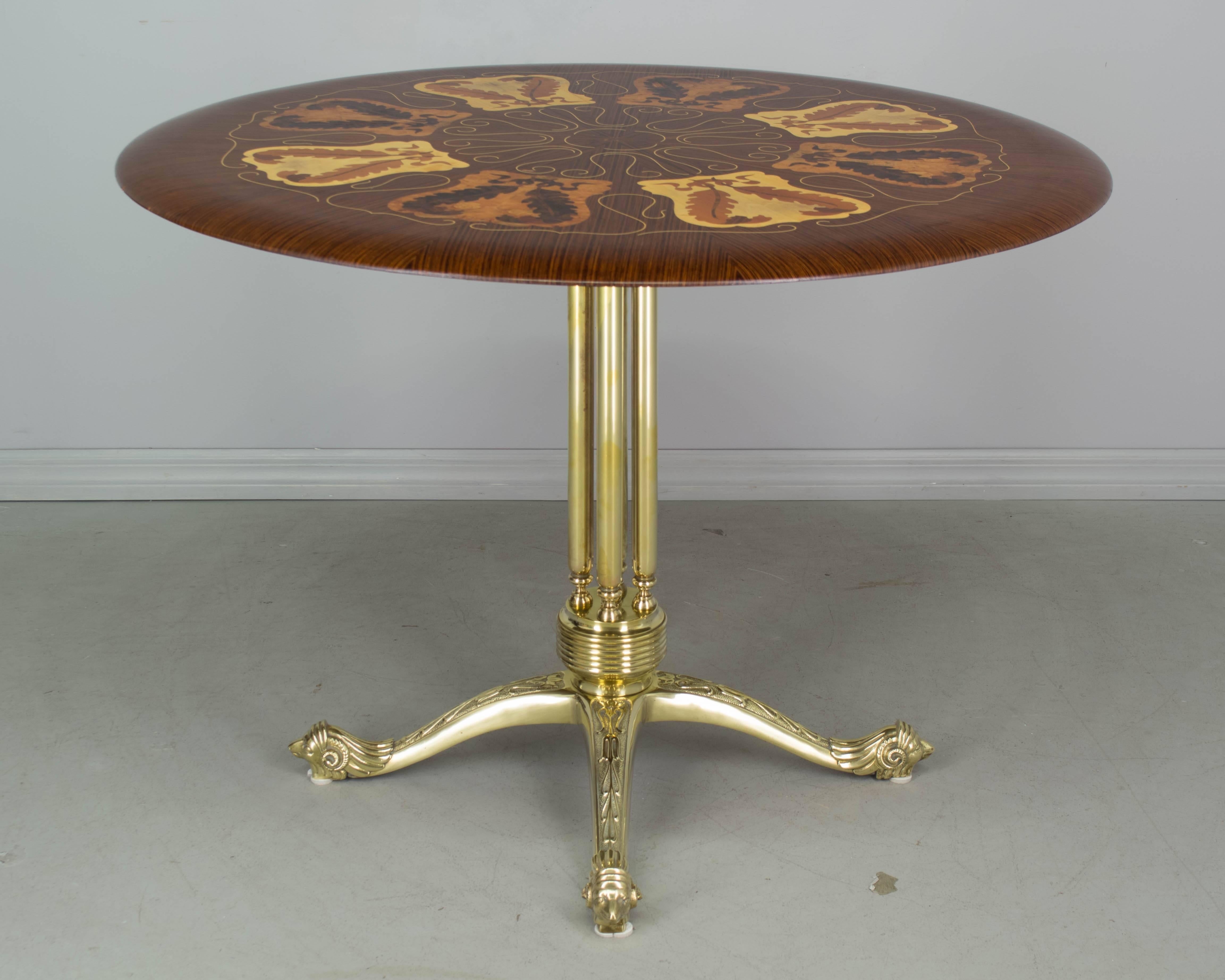 Italian Mid-Century circular table with brass base and marquetry top. Pedestal base with four central tubular columns. Solid brass legs with decorative embossed designs ending in sculptural ram's heads. Top is inlaid with exotic woods and has high