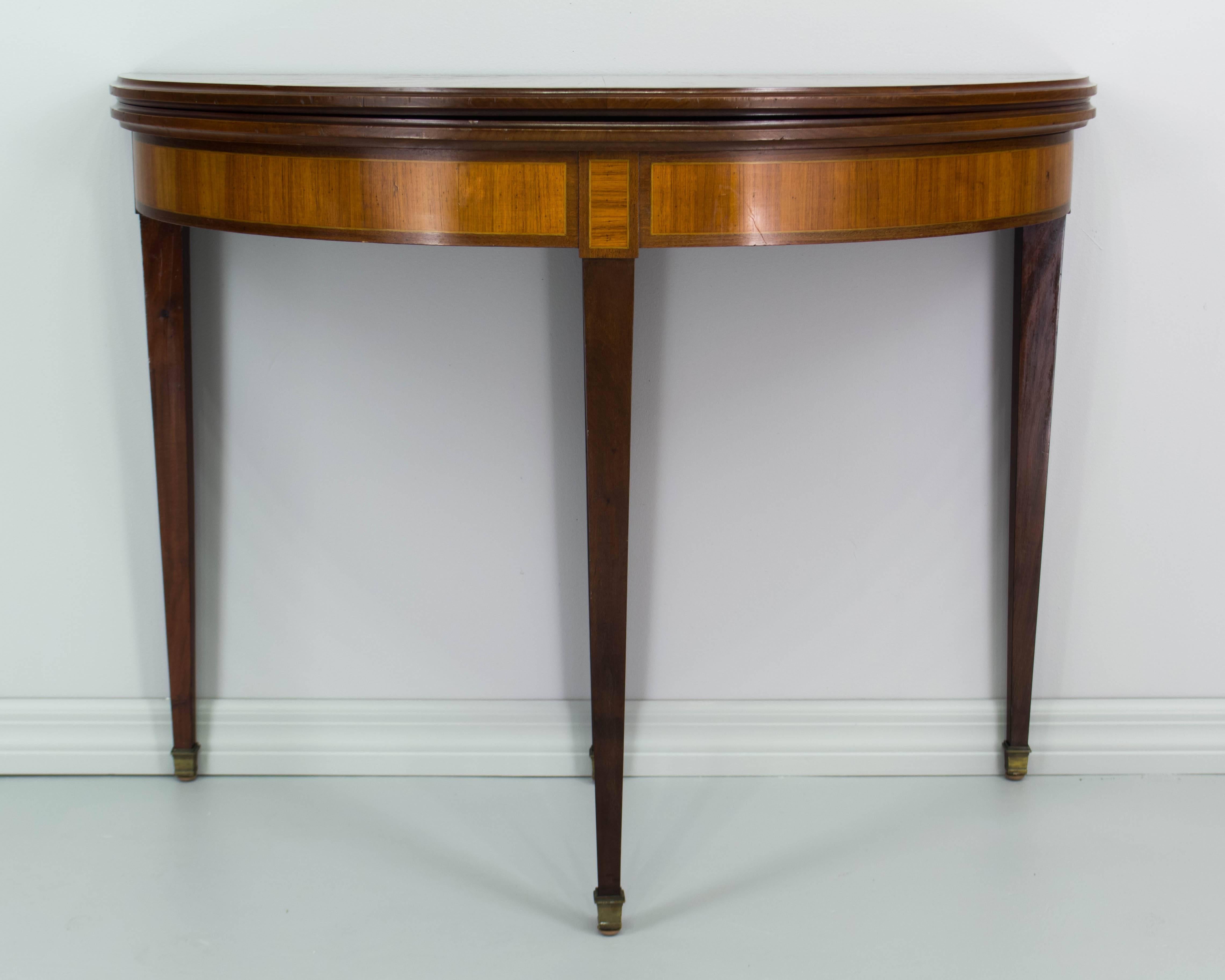 A fine French Louis XVI style demilune console that opens to a game table. Marquetry top is inlaid with veneers of mahogany and walnut. Four tapered legs ending in brass sabots. Top hinges open and rests on center leg which pulls out to reveal