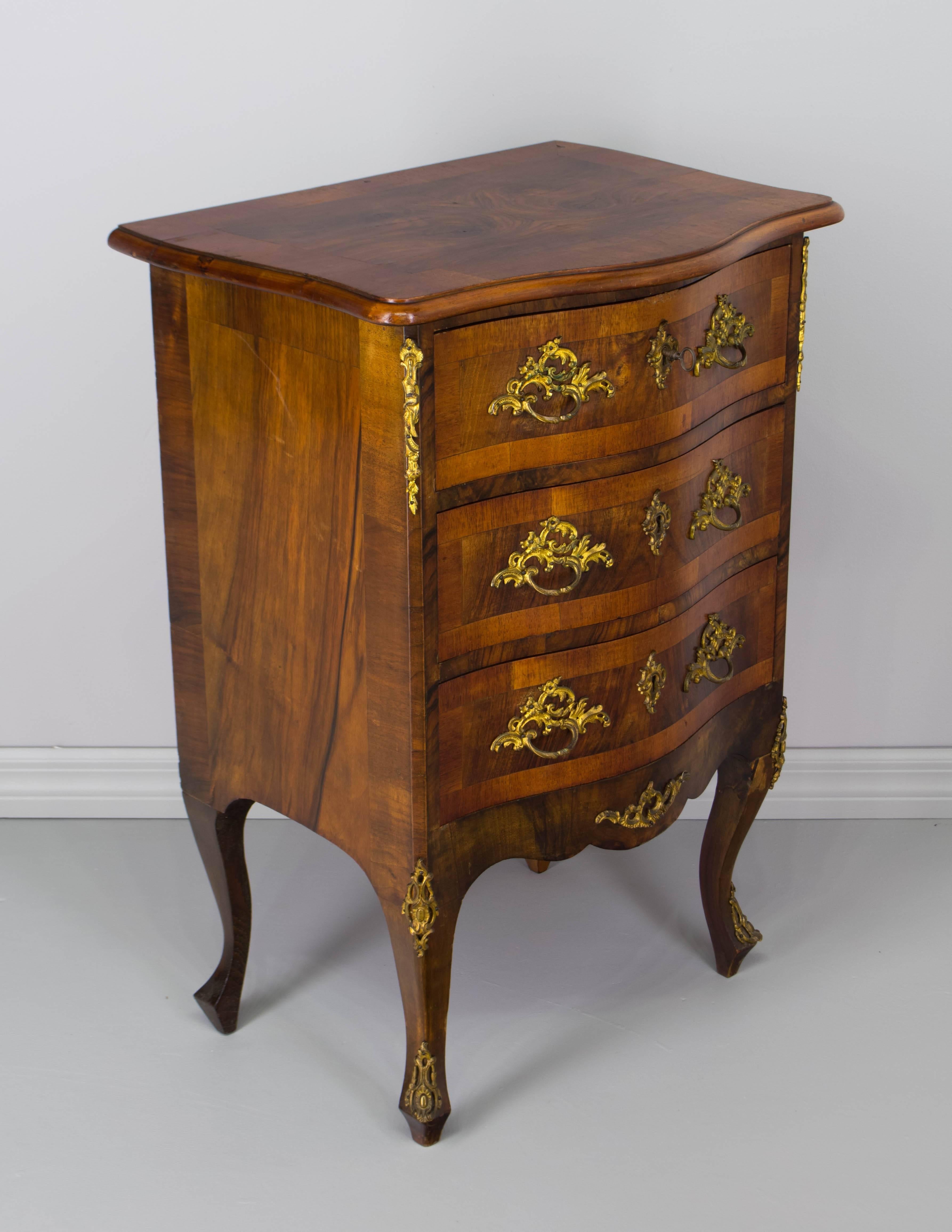 A small Louis XV style commode with serpentine front and three dovetailed drawers. Beautiful veneer of walnut with solid oak as secondary wood. Original bronze hardware. Three working locks with one key. A nice size for use as a nightstand. 
More