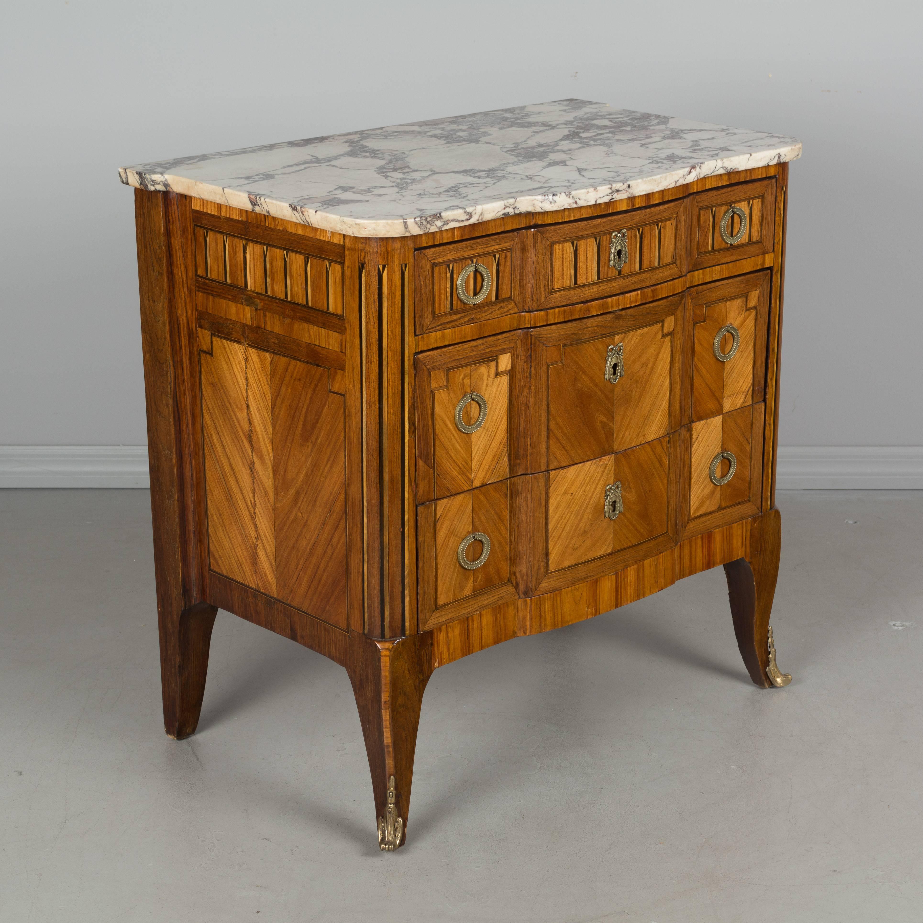 A 19th century French Louis XV style three-drawer marquetry commode inlaid with veneers of mahogany and tulipwood. Original marble top. Brass hardware and brass sabots on front legs. French polish finish. Locks are present, but there are no keys.
