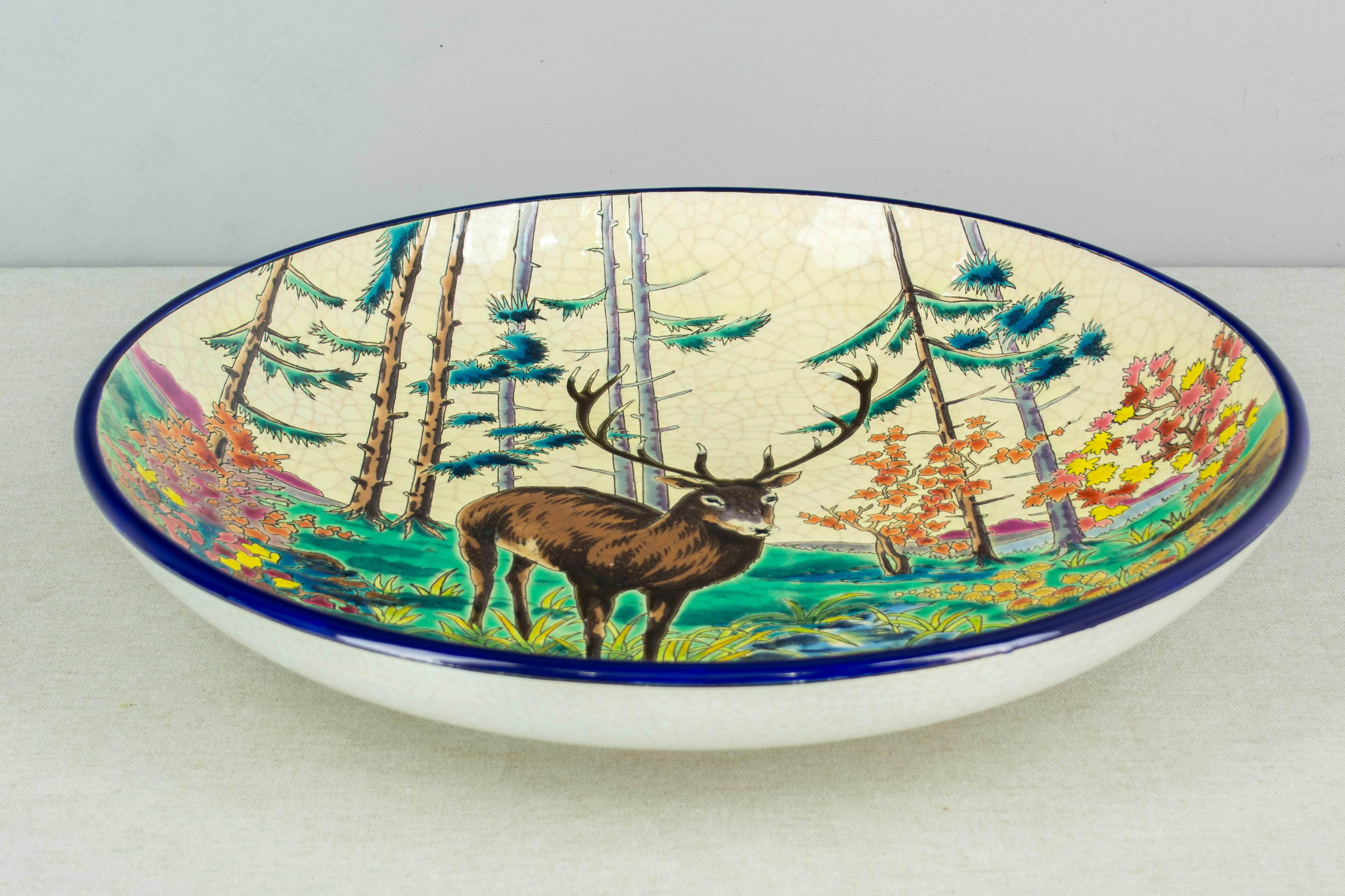Large decorative French Longwy pottery deep wall charger, enamelled with a buck near a stream surrounded by colorful foliage. Craquelle white ground and cobalt blue rim. Beautiful vivid color and unusual large size. Designed by Paul Mignon and