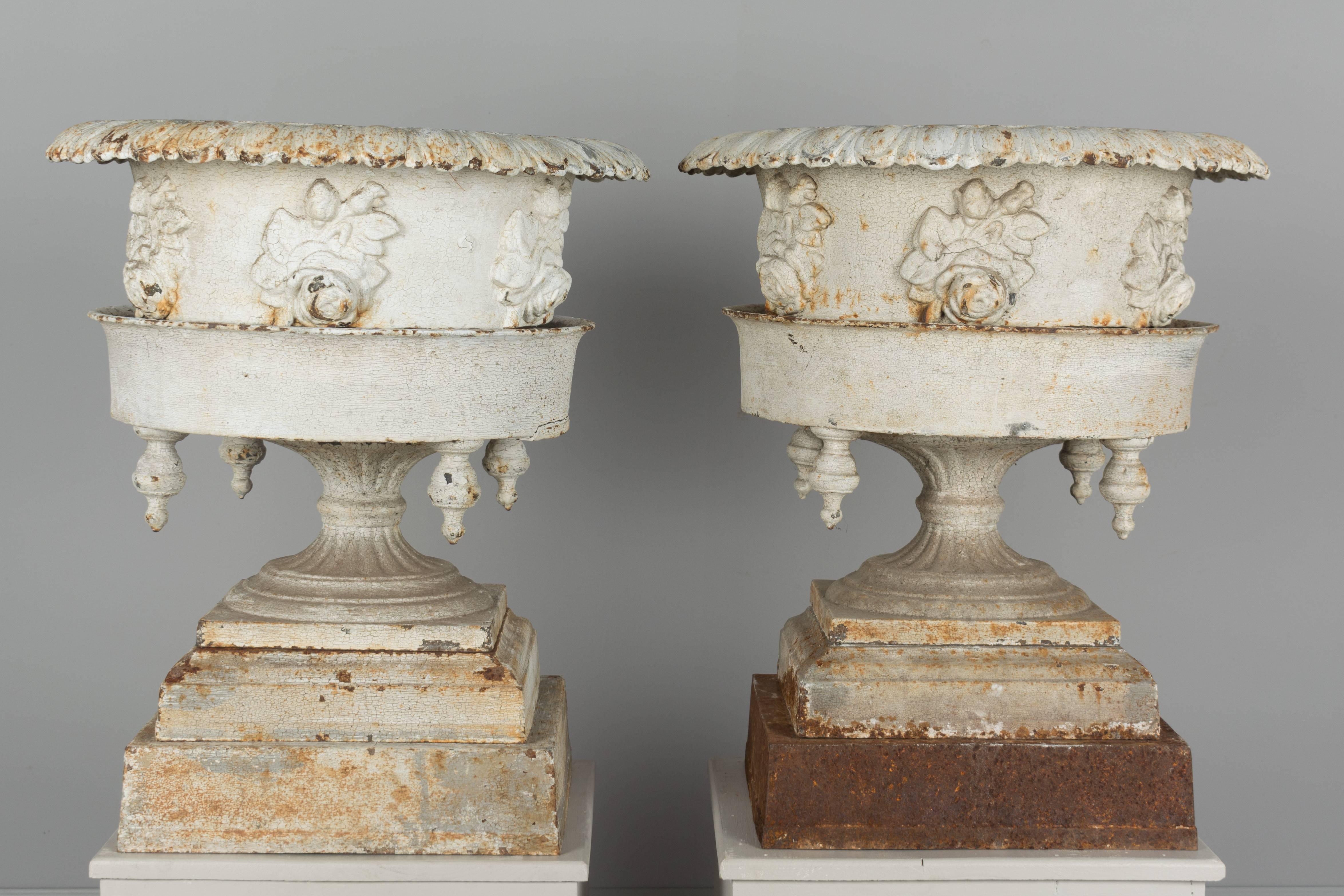 Pair of American cast iron urns with original old white painted patina. Found in northern New York State. These planters have an unusual form with a large decorative urn adorned with roses that sits inside a wider base with four finial details, all