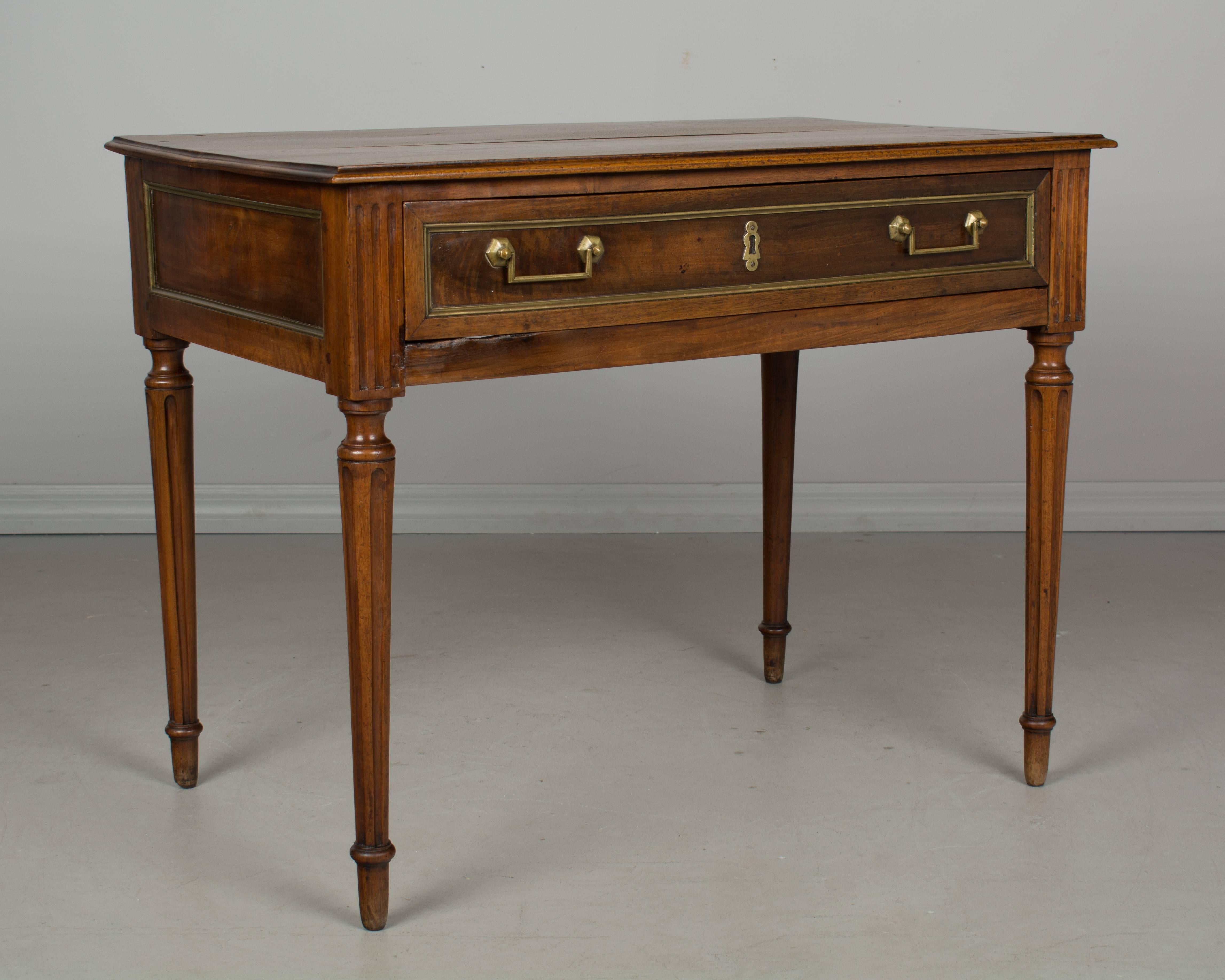 A 19th century French Louis XVI style writing table or side table made of walnut with brass inlaid trim. Large dovetailed drawer with brass pulls. Keyhole but no lock. Slender turned fluted legs. Waxed finish. All original. Clearance of 21 inches.
