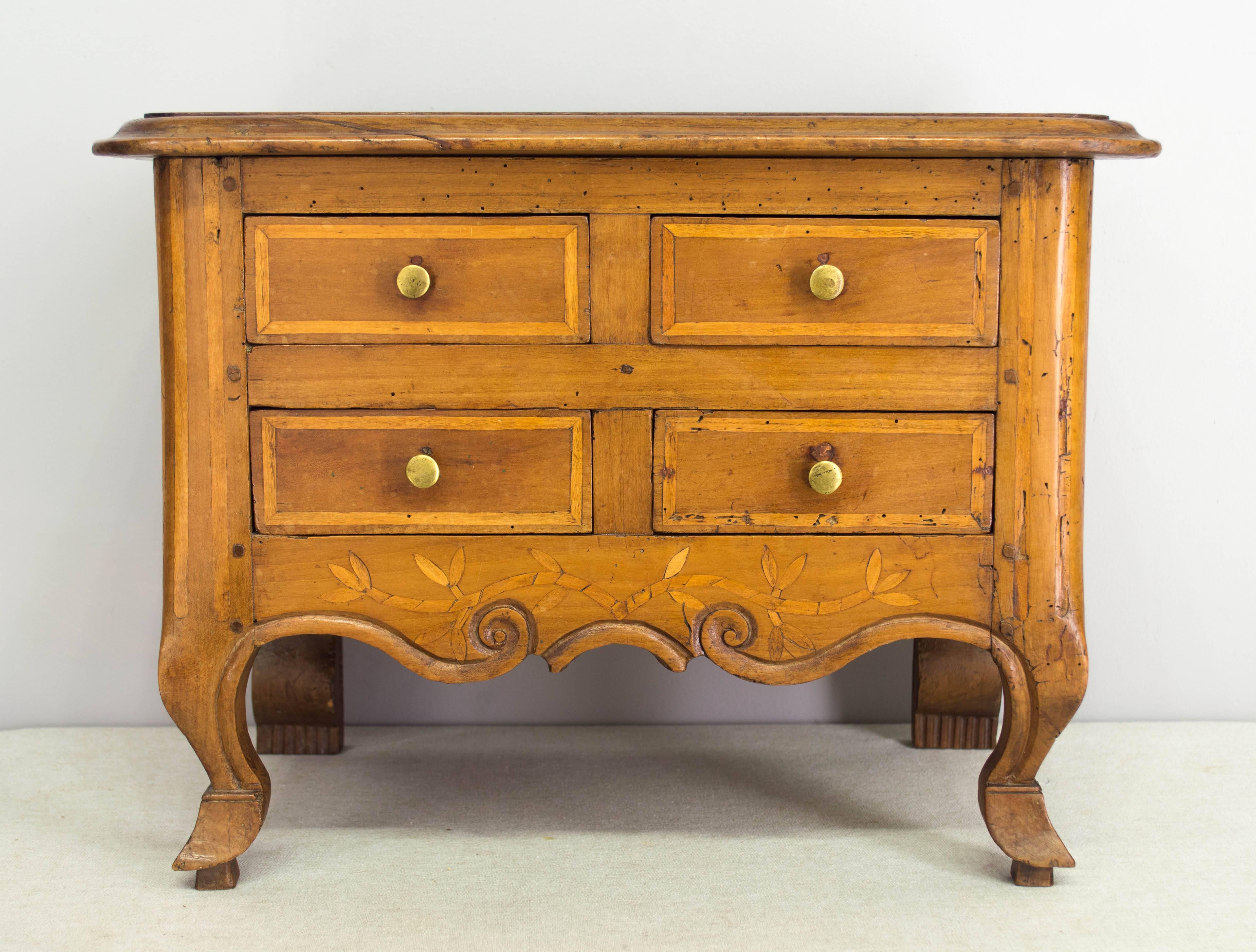 An early 19th century Louis XV style miniature commode, made of cherry with four dovetailed drawers. Nice details to this little chest including curved front corners and legs ending in hoof feet. Carved apron with inlaid vine decoration. Waxed