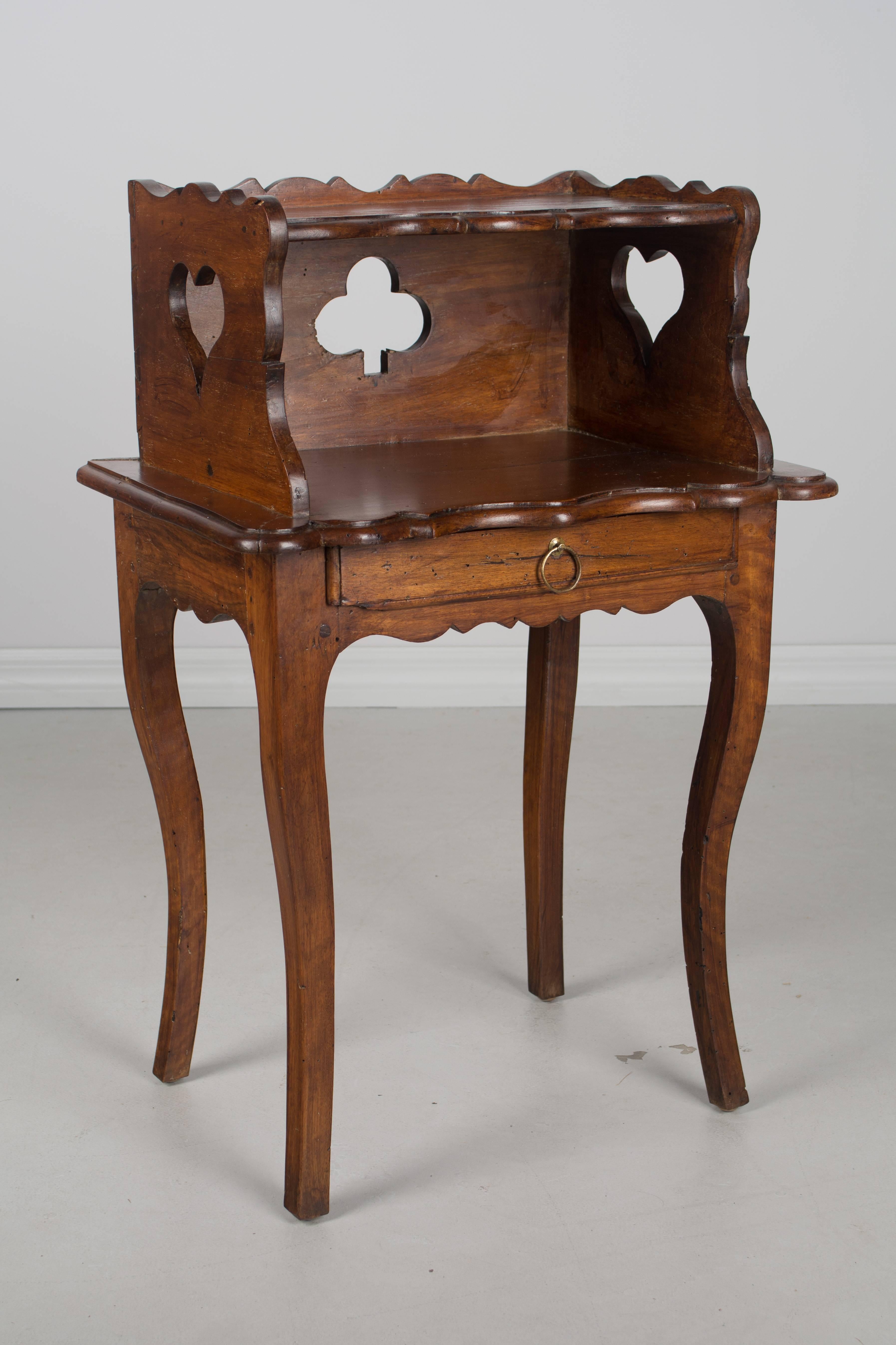 A late 18th century Louis XV country French side table or nightstand made of solid walnut, with pierced cloverleaf on back and pierced hearts on the sides. Scalloped apron and gallery surrounding the upper shelf. Small dovetailed drawer with brass