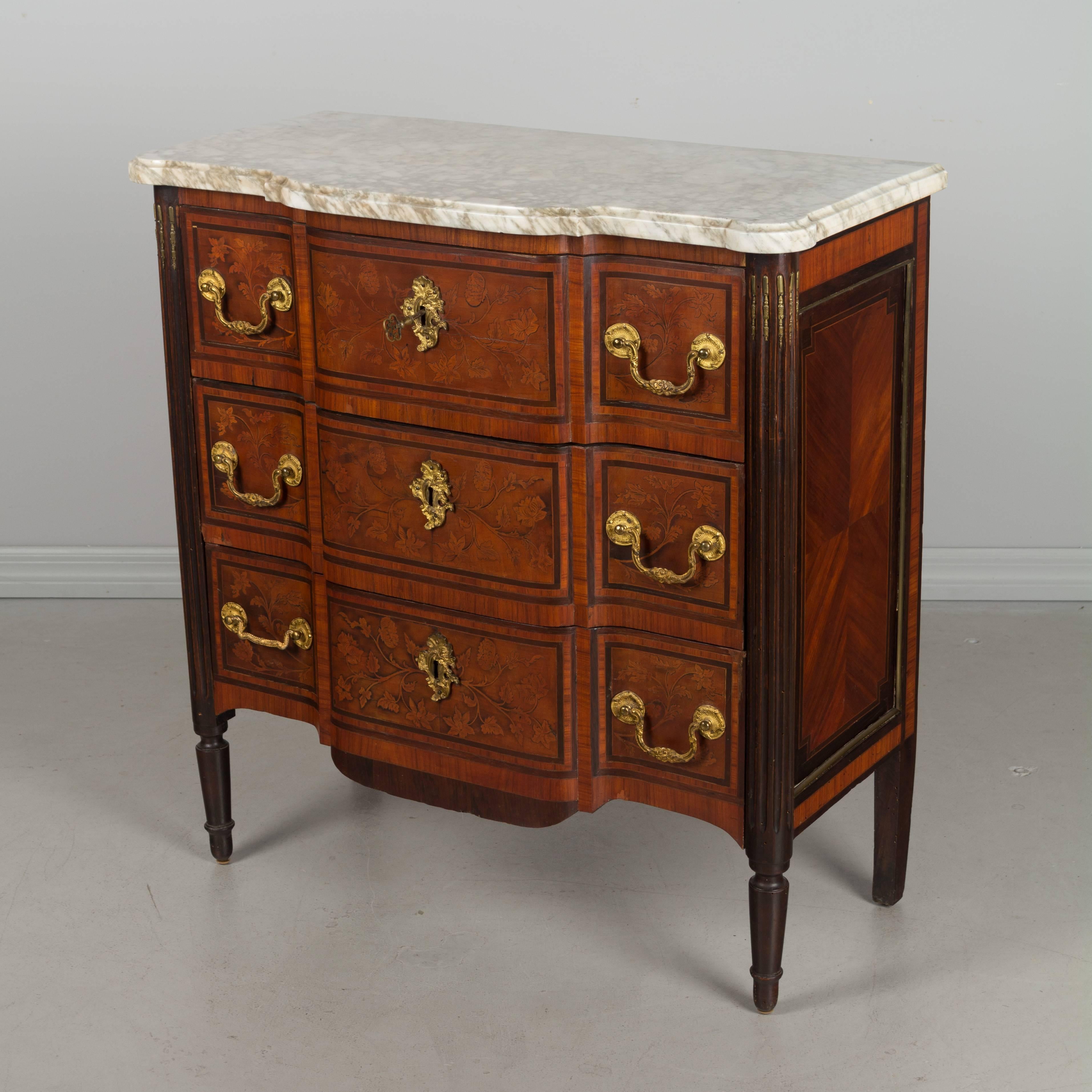 An early 20th century French Louis XVI style marquetry commode with inlaid veneer of mahogany and brass decoration. Original white veined marble top. Three drawers with brass pulls. The bottom left drawer pull does not match the others. Locks in