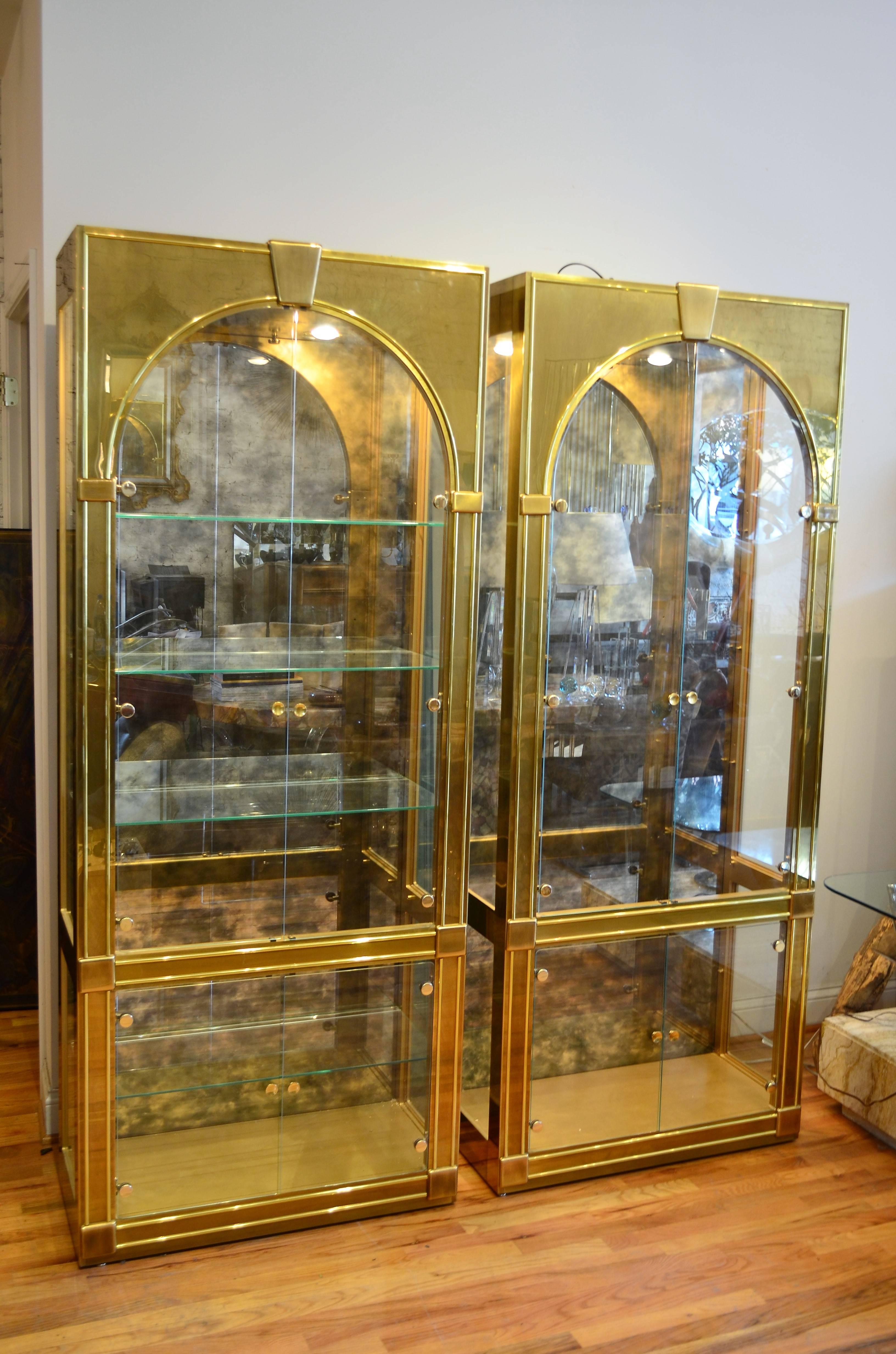 Pair of brass Mastercraft Palladian style vitrines with antiqued smoked mirrors backs.
Each cabinet is lite by two dimmer controlled interior lights
and has five adjustable glass shelves .