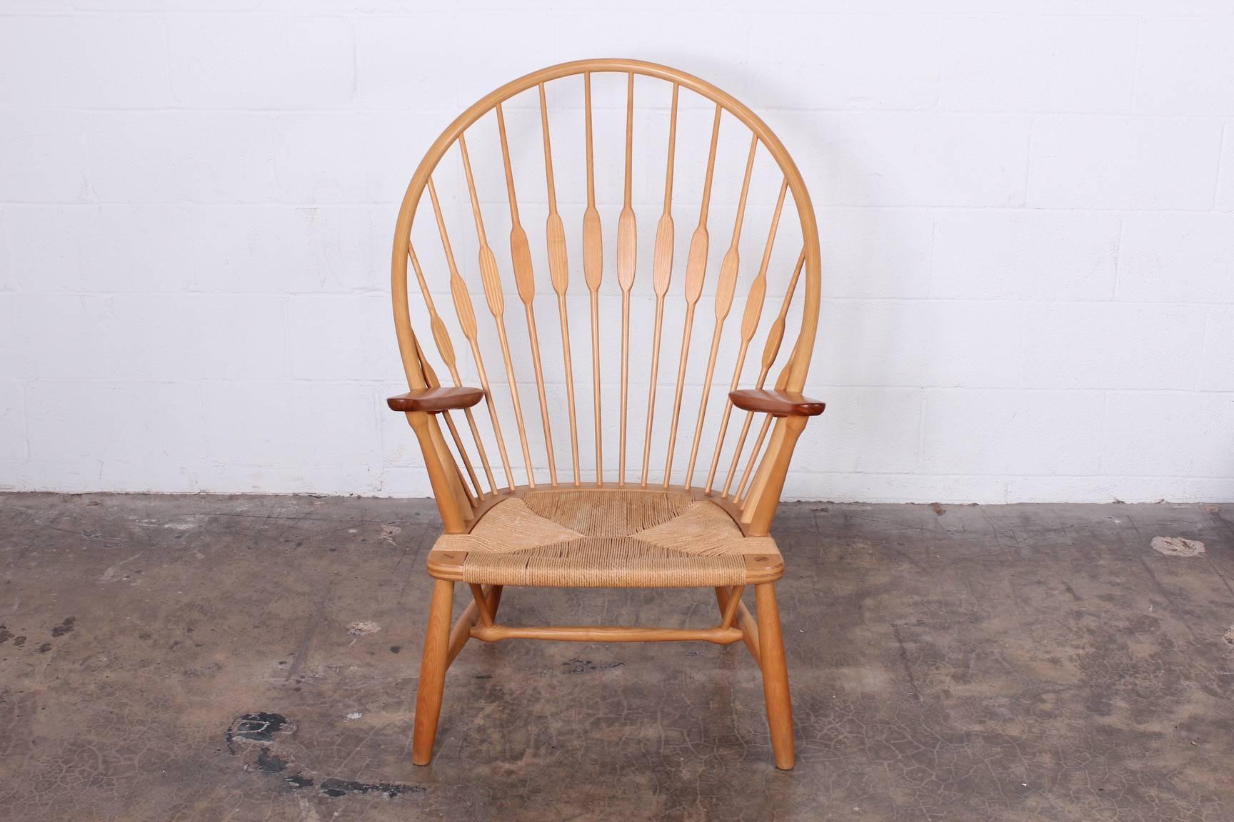 The 'Peacock' chair was designed by Hans Wegner in 1947 and manufactured by Johannes Hansen, Denmark. It features a solid ash frame with a rounded high back, contrasting teak arms, and a woven paper cord seat.