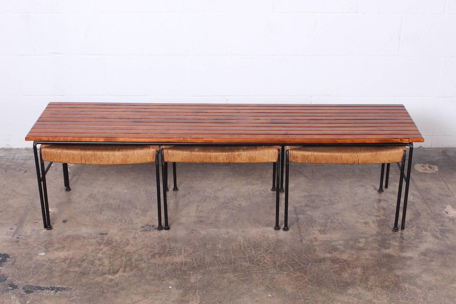 A slatted maple bench with three rush stools designed to store underneath by Arthur Umanoff for Raymor.