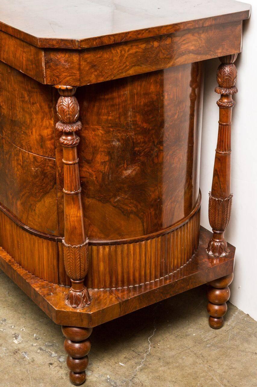 Wonderful, two drawer, oval, burl veneered commode with fluted band, nestled between clipped corner plinths and four, foliage embellished column supports. The whole on tapered, bubble-style feet.