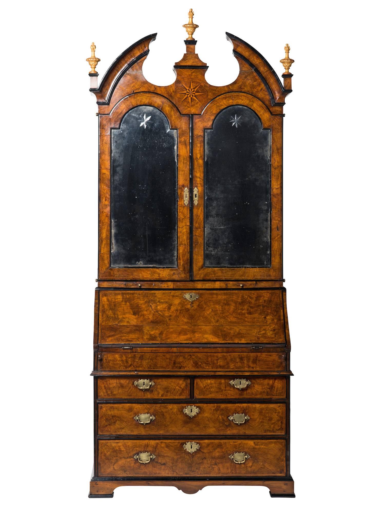 The upper part having a swan’s neck pediment centered by an inlaid star medallion and surmounted by gilt finials, over a pair of mirror-fronted doors opening to a fully fitted interior of small drawers, folio slides, pigeon holes and secret