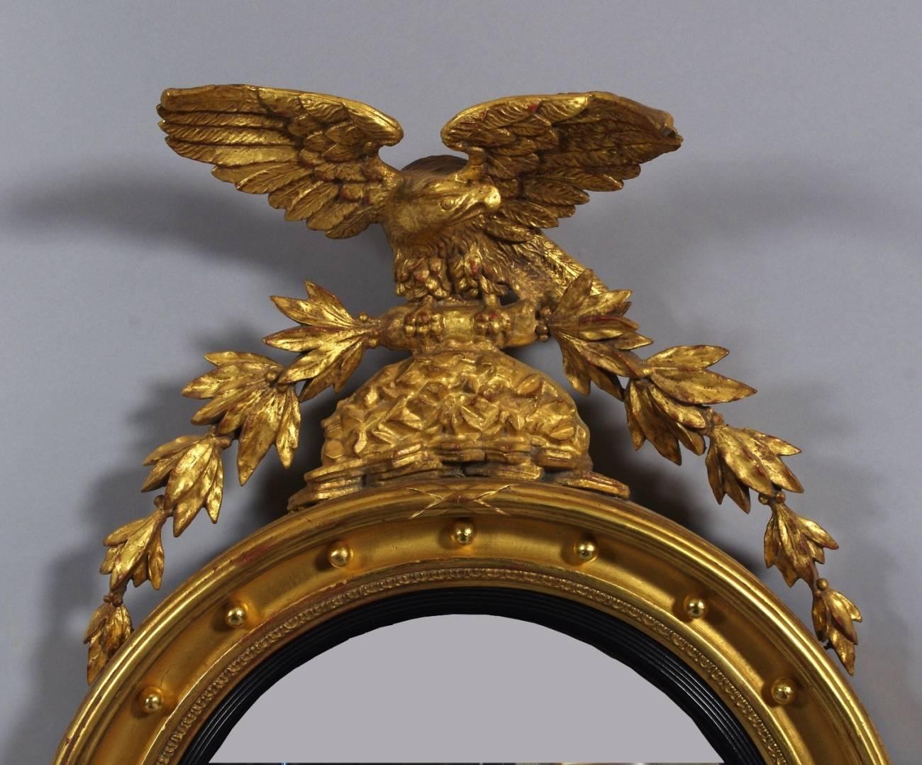 Regency carved and giltwood convex mirror, the crest surmounted by an eagle with out-stretched wings on a rocky outcrop, a bundle of laurel branches in its claws, the frame with gilt balls and ebonized liner surrounding the convex mirror.