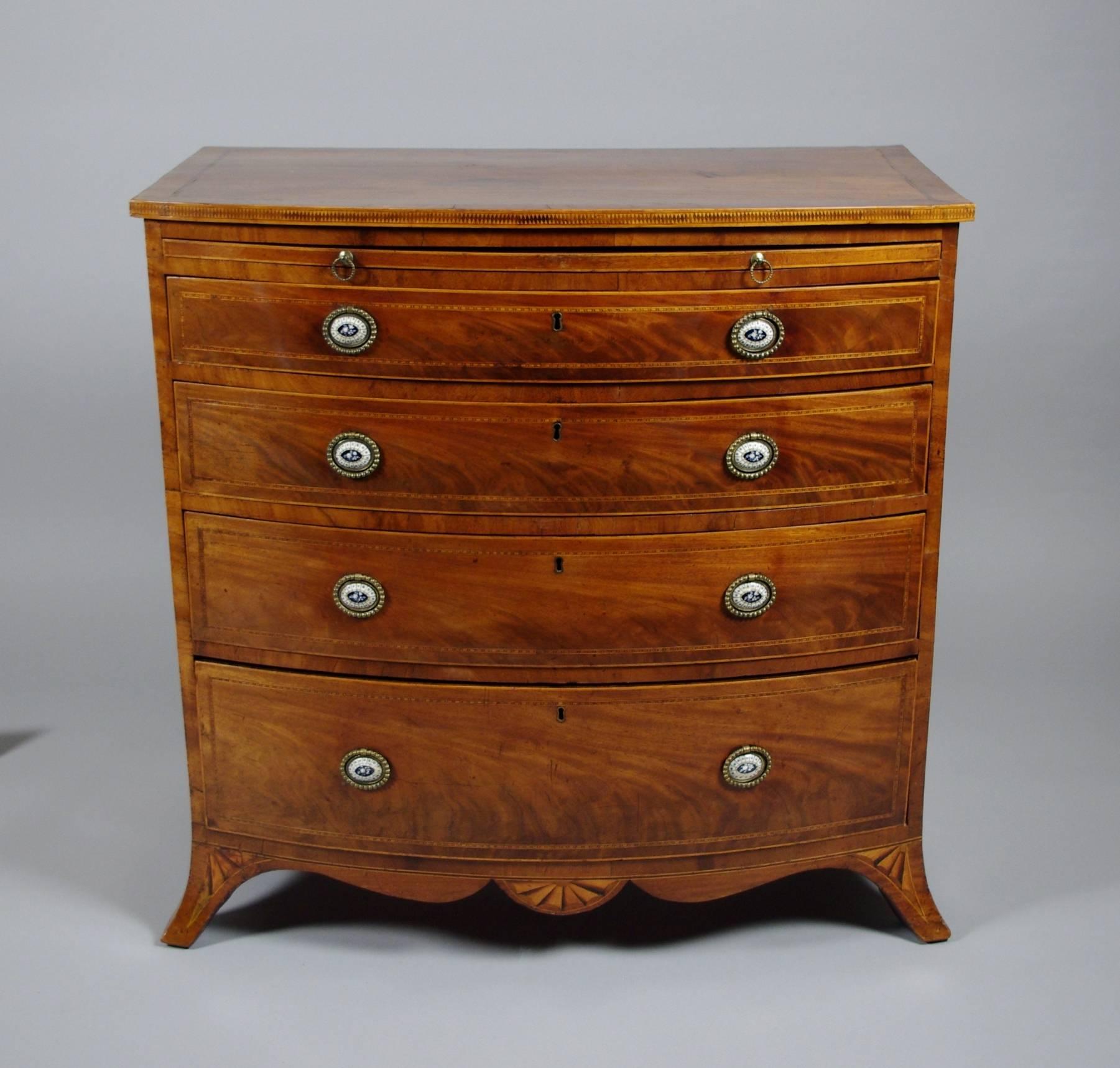 George III mahogany bow front chest of four long drawers, the banded top with string inlay and harlequin edge over the pullout brushing slide; the four graduated drawers with well figured veneer and chevron stringing having the original enamel