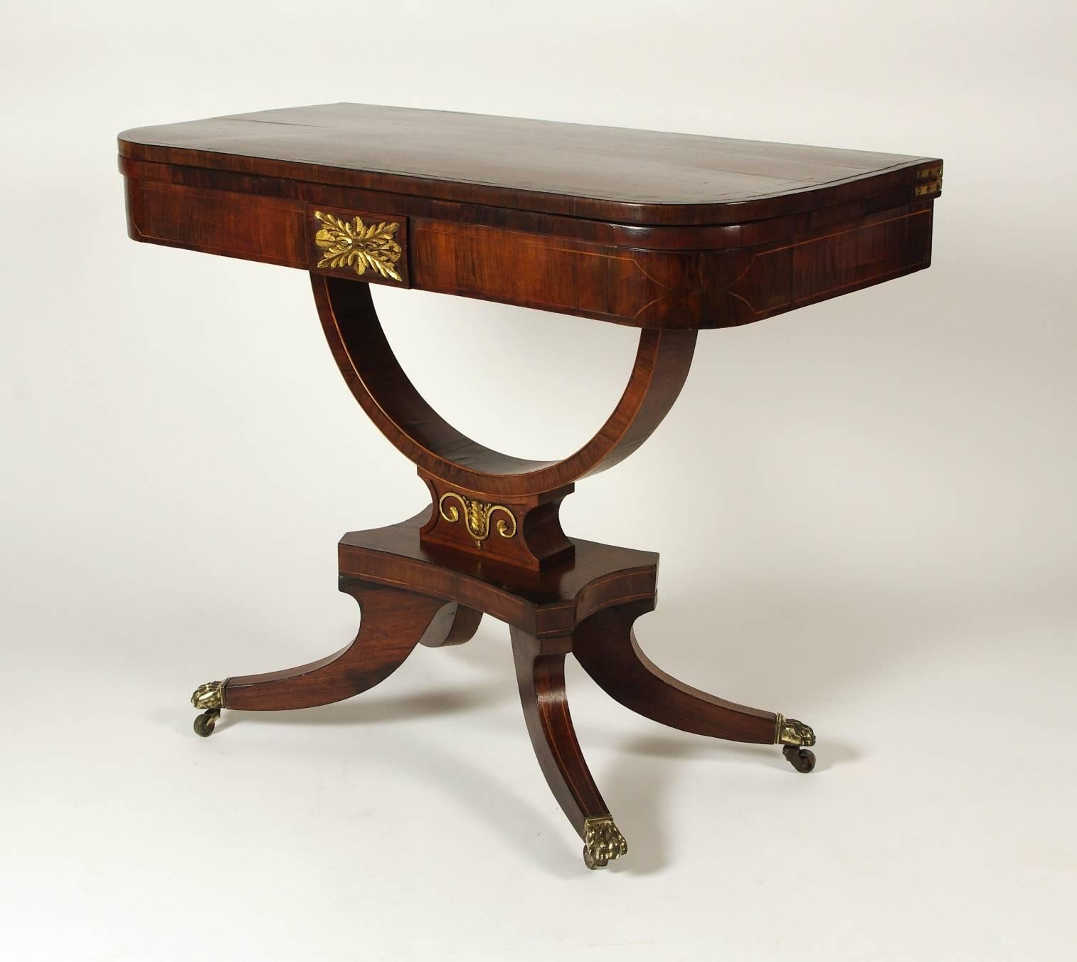 Regency rosewood fold over card table, the rectangular top with rare palm-wood crossbanding opening to a replaced tooled black leather interior; the apron with string inlay centered by a leaf carved and gilded medallion on a stylish arched support