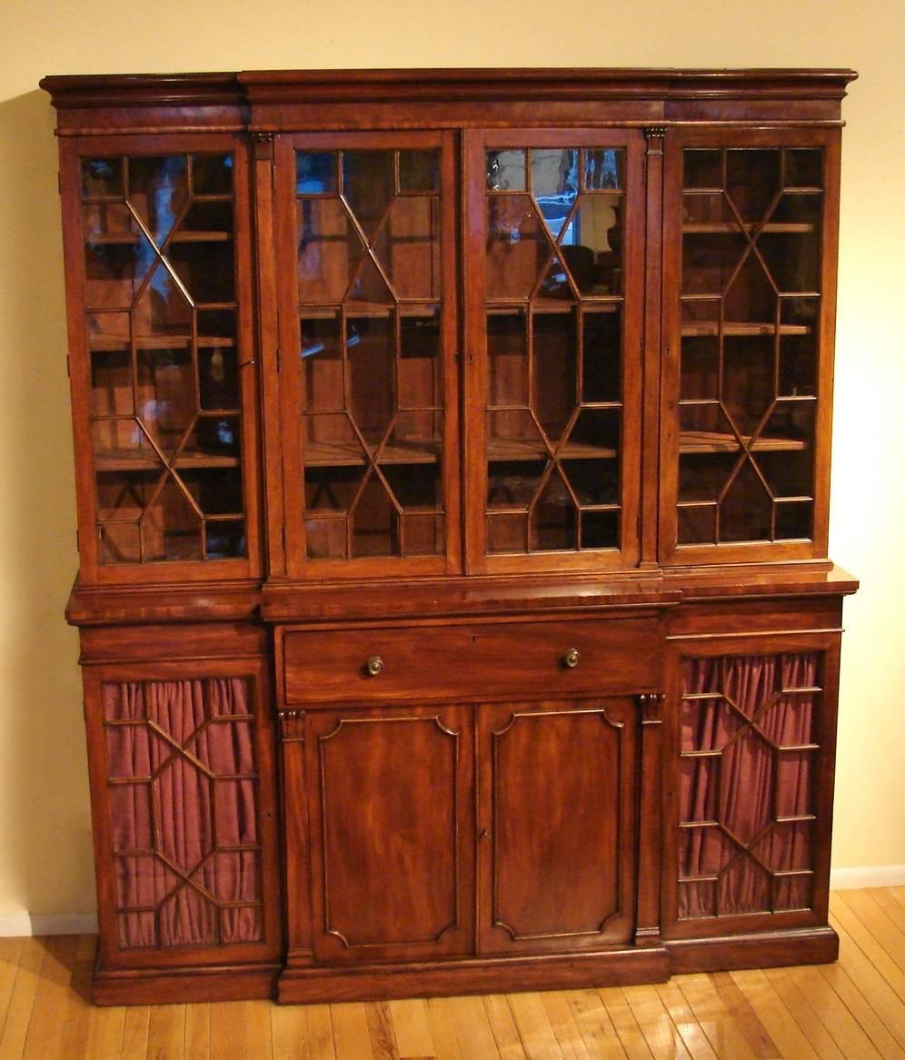 A late Regency period mahogany breakfront bookcase of compact size, the astrgal glazed upper section with four cabinet doors above a fitted secretaire above a pair of solid central cabinet door flanked by opposing glazed doors supported on a plinth