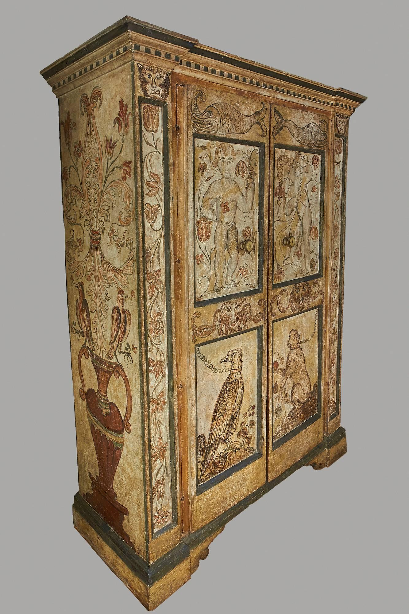 A wonderful cream and grey painted Italian Baroque period cupboard, the blue banded outset pediment with a painted trompe l'oeil dentil, over twin paneled doors headed by opposing ferocious fish above figures of boys entwined in floral vines, the