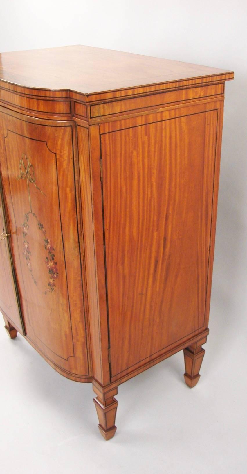 A good quality English Sheraton style satinwood line inlaid and paint decorated music cabinet, the case decorated with polychrome bows and floral garlands, the top with floral painted border, the doors opening to a single shaped shelf all raised on