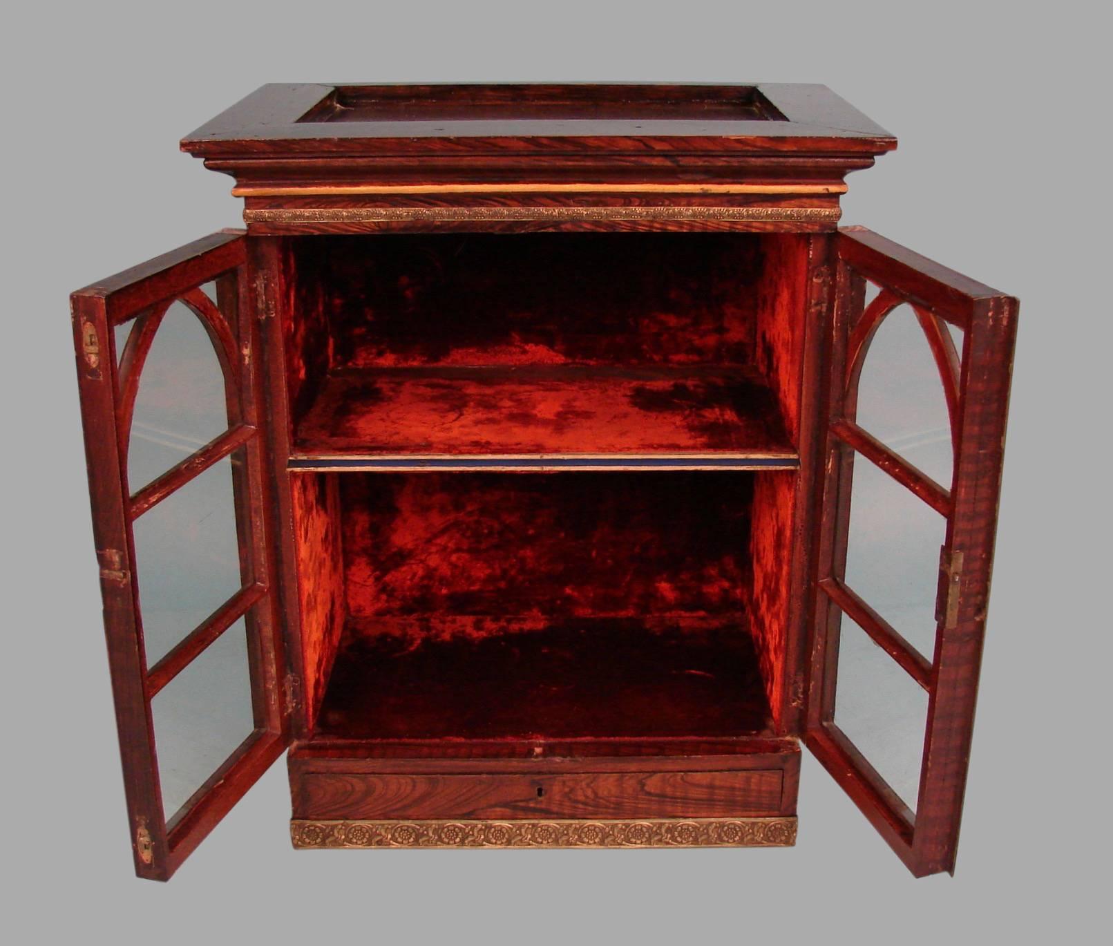 An English 19th century grained-painted table cabinet, the cornice resting above two Gothic style astragal glazed doors concealing a velvet lined interior with one shelf, the base fitted with a single narrow drawer. The piece is banded with gilt
