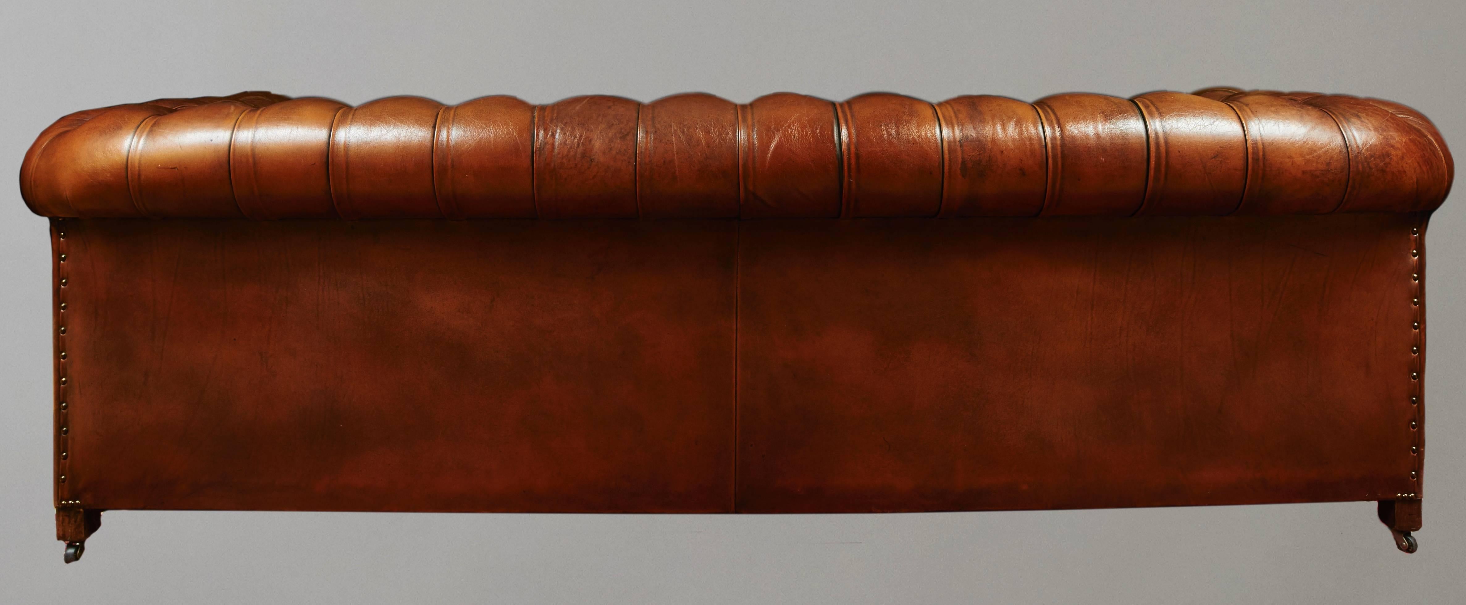 A good quality English or American tufted leather sofa upholstered in chestnut colored hide in traditional style with nailhead trim and rolled arms, resting on bun feet, 20th century.