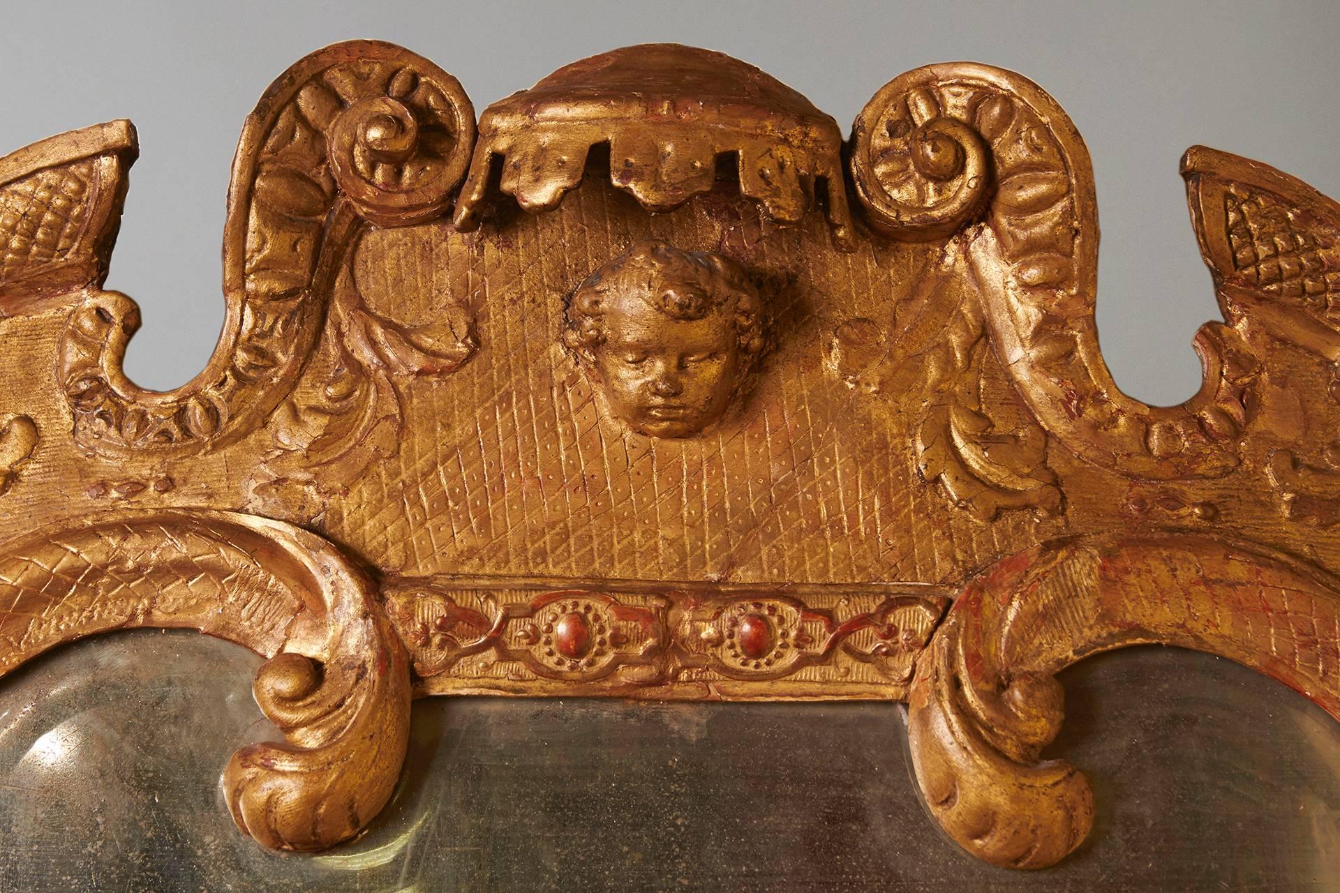A George II carved giltwood mirror, the arched surround with foliate details  centering a female mask, retaining its original 2 part beveled glass plate, the burnished relief decorated sides terminating in scrolls. Circa 1730.