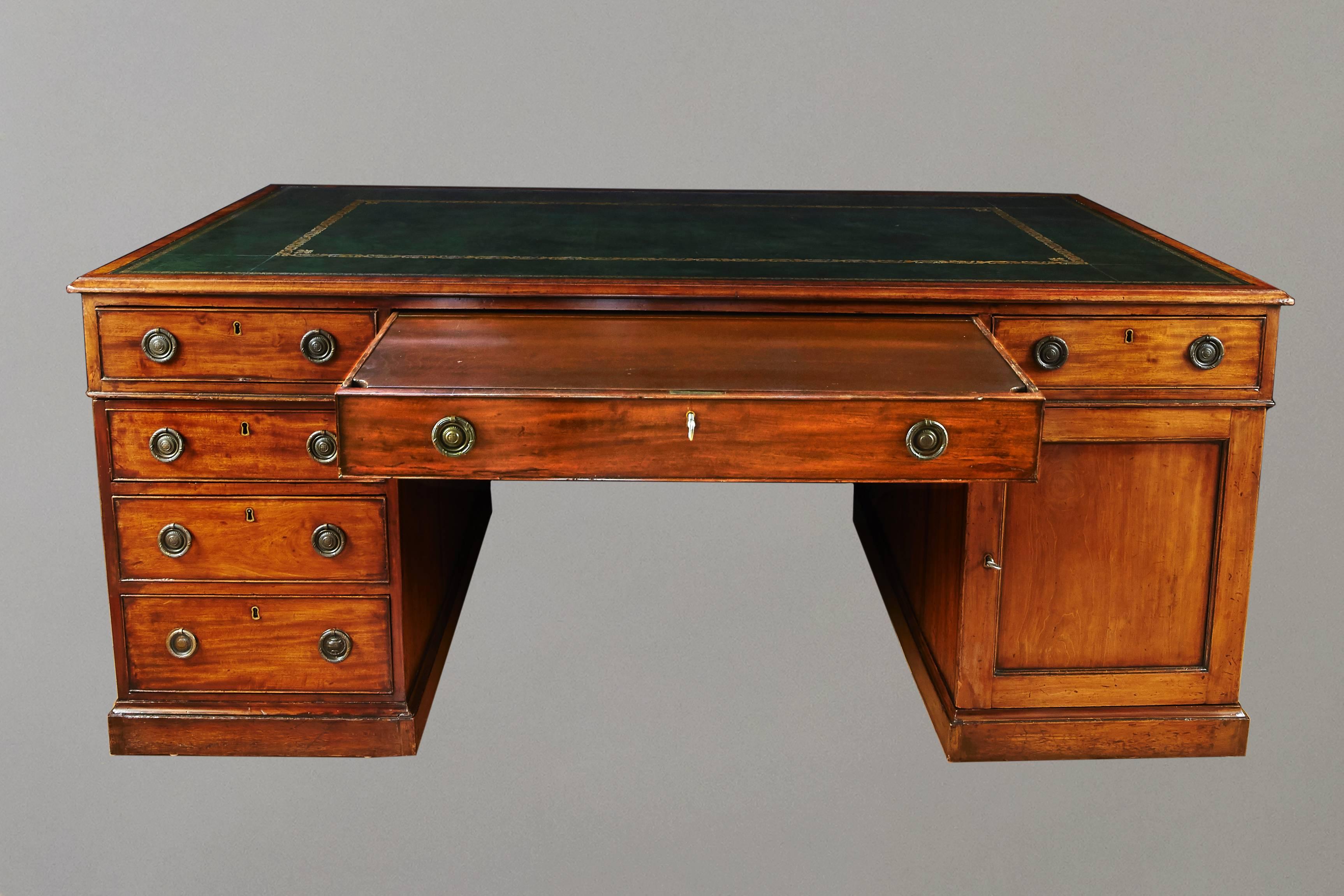 A George III style mahogany partners' desk, the green gilt-tooled leather top over three frieze drawers above three graduated drawers on one side and a cupboard on the other, all resting on a plinth base. Reverse identically configured, 19th