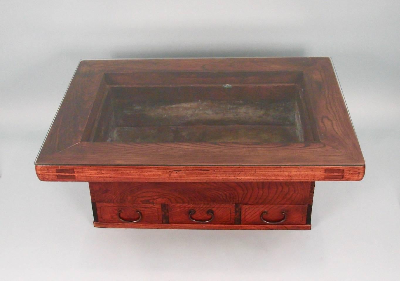 A Japanese keyaki wood hibachi, the copper inset liner framed by a thick surround resting on a well-figured keyaki base with three small drawers and side carry handles. Comes with a later fitted glass top.