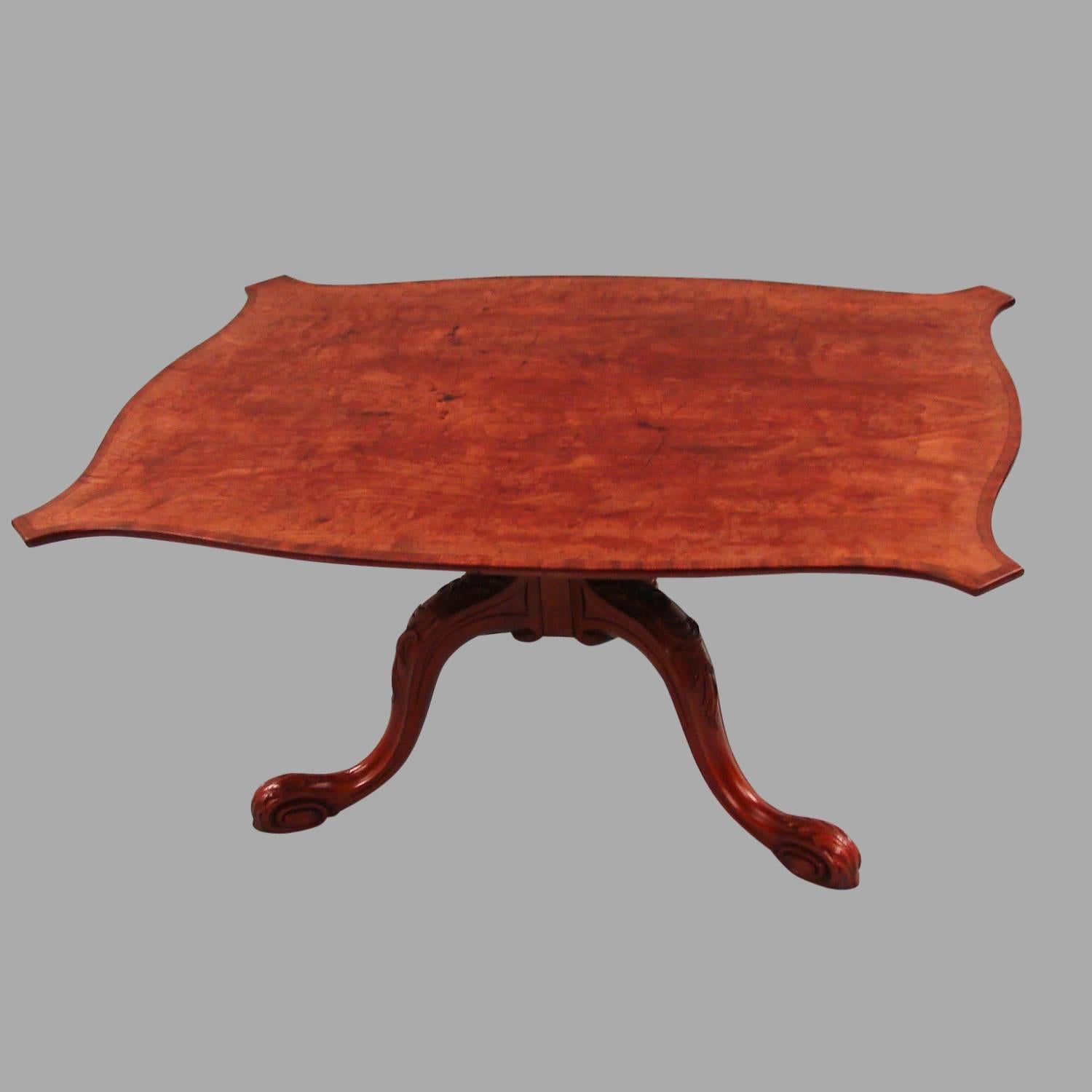 A George III inlaid mahogany tilt-top table, the serpentine form crossbanded top supported on a carved tripod base ending in pad feet. Reduced in height.
