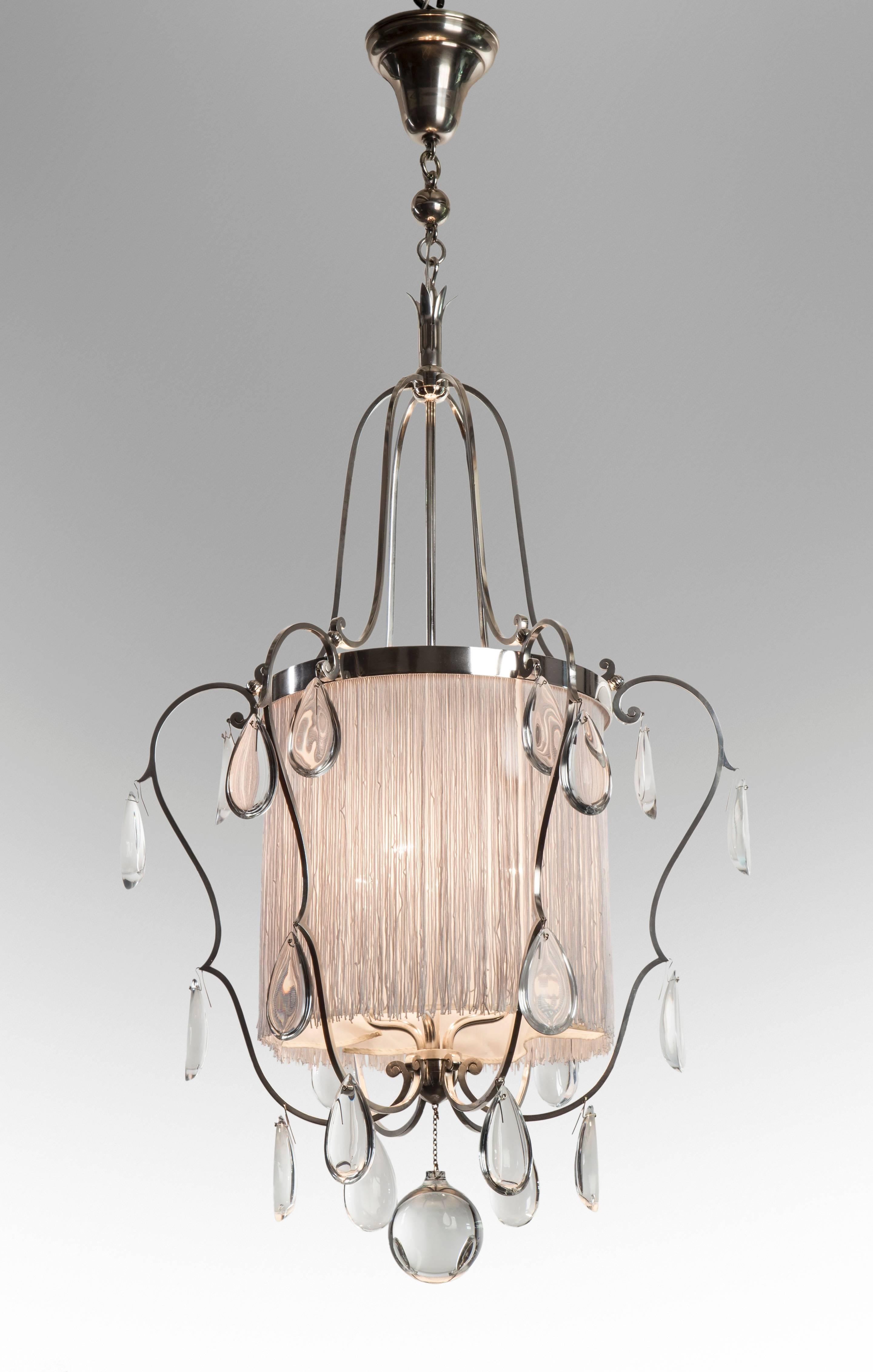 A graceful and exuberant chandelier of the highest quality. The cylindrical body dressed with silk tassels, fitted with six inward scrolls hung with clear faceted pear-shaped crystals, ending in a large sphere and the silver lacquered.