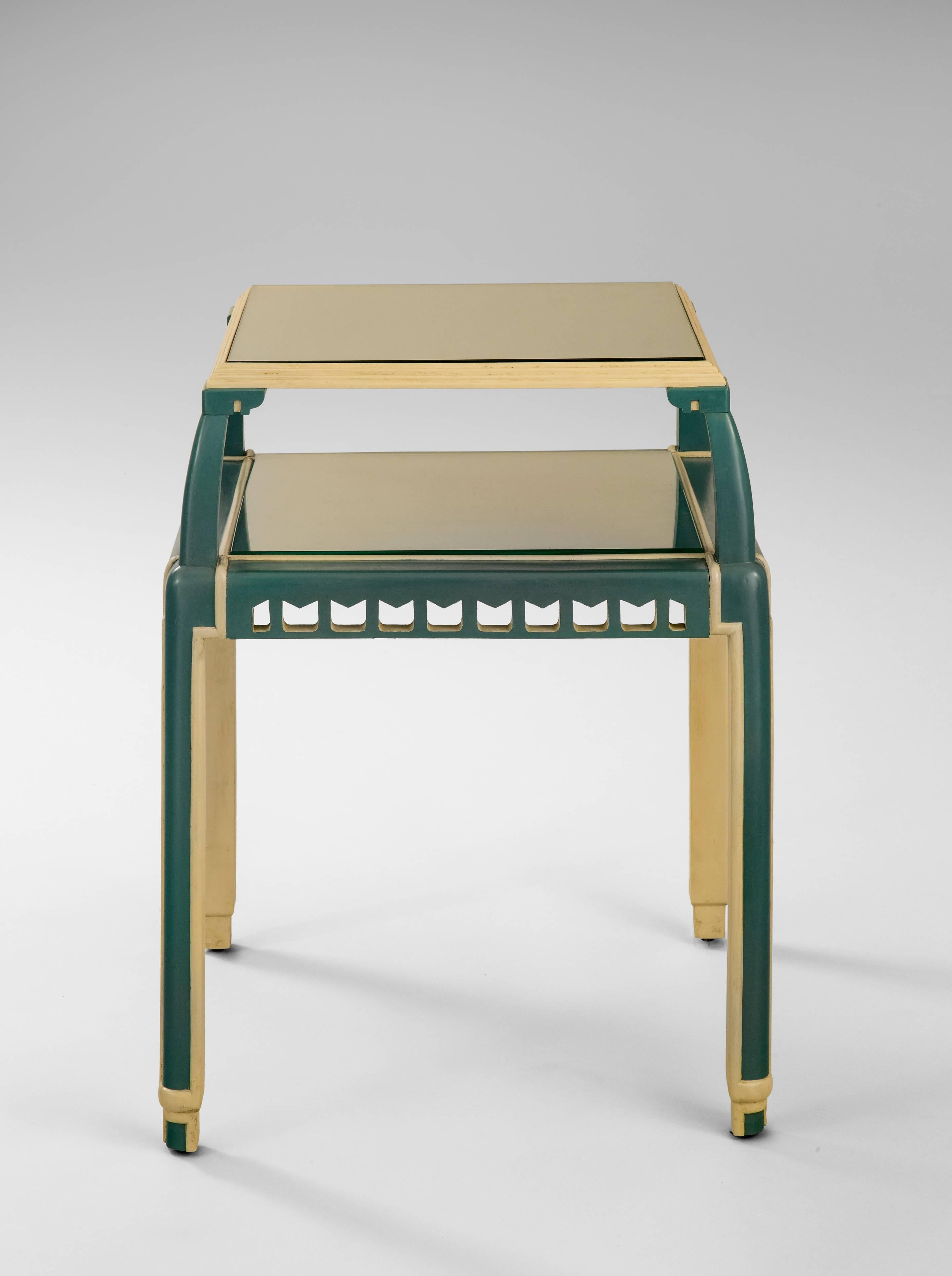 A charming table of crisp design plus a shelf for an extra display area. The square top with stepped molded border, raised on stylized capitals, above a lower shelf with pierced apron, on four molded legs, terminating in recessed demilune feet. 