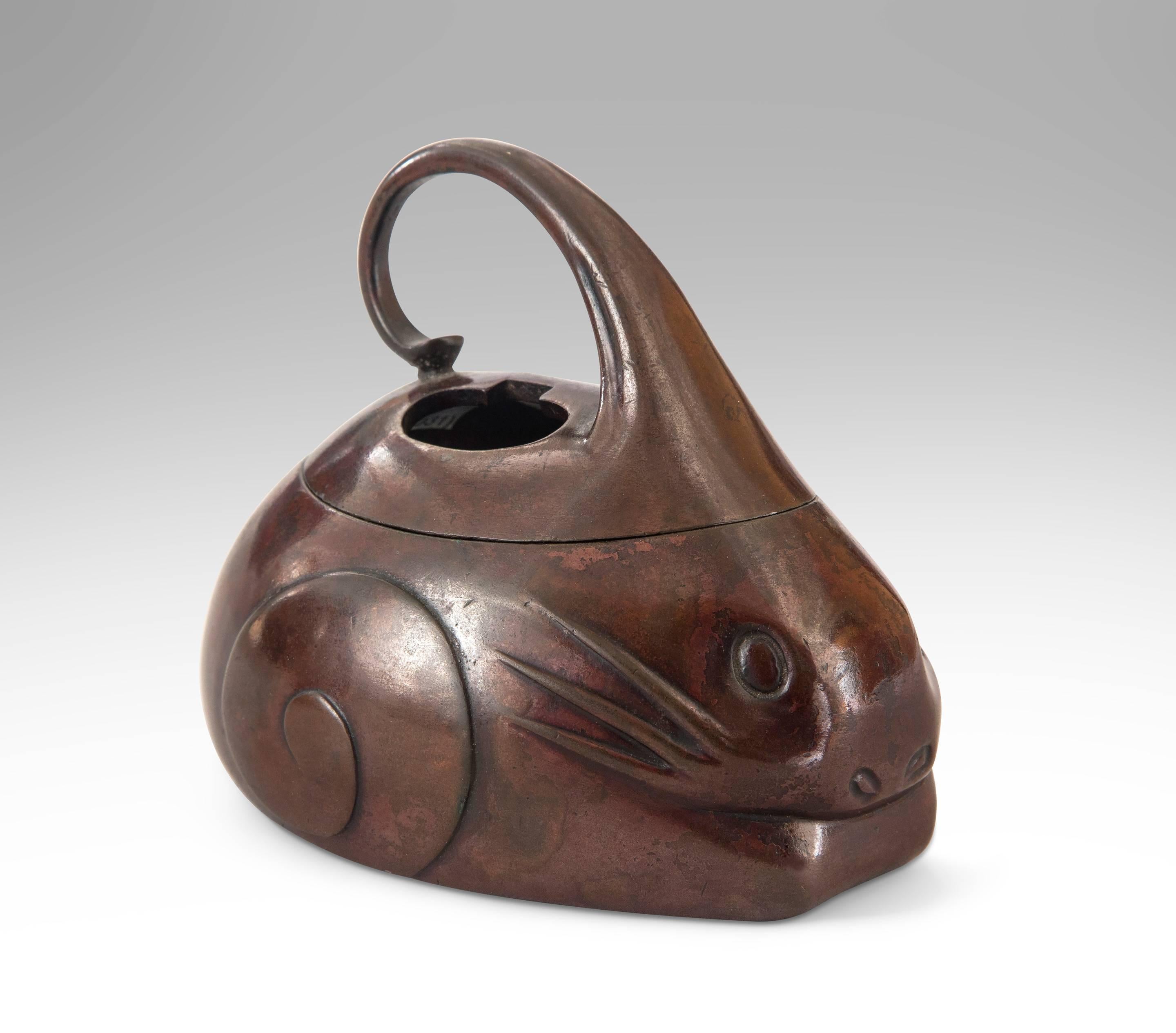A charming and rare model with moderne style. In the form of a crouching rabbit with incised eyes and low relief whiskers, the ears formed into a scroll handle which opens the pierced back to a hollow interior.

With a Tomobako (original wooden