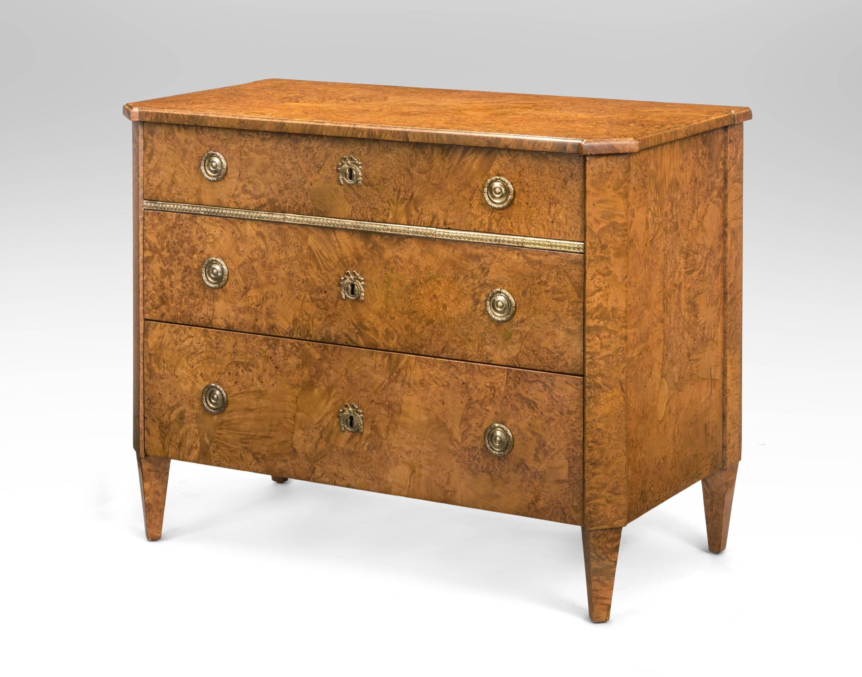 A hard to find Neoclassical commode of beautiful golden colored burlwood. Perfect for a contemporary or eclectic interior. The molded rectangular top with canted corners and break-front sides above one frieze drawer and two lower drawers graduating