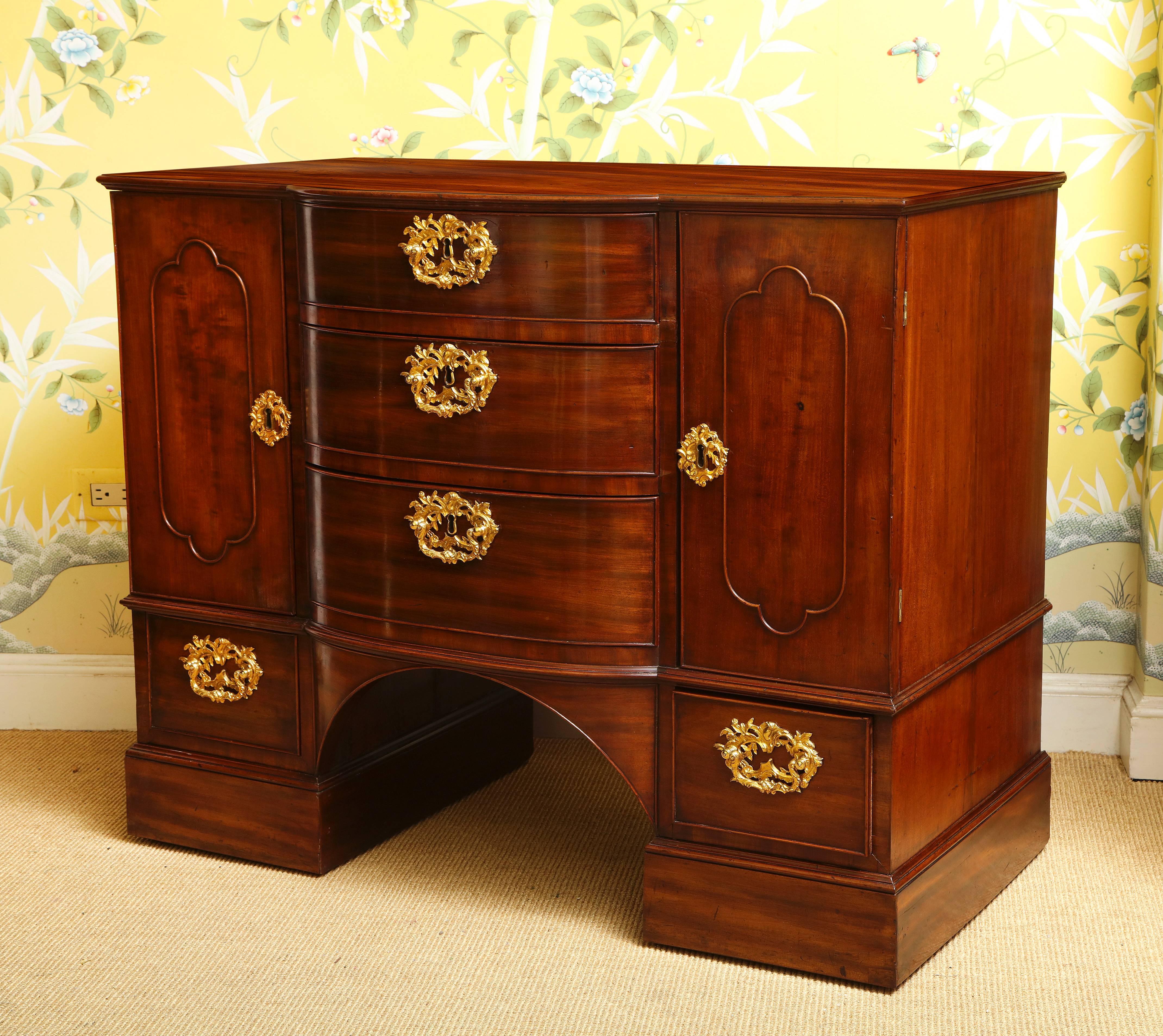 Very fine George III ormolu-mounted mahogany commode, having a moulded rectangular bowed center above three graduated cockbeaded drawers above the arched center and flanked on either side by a shaped paneled door and short drawer mounted with gilt