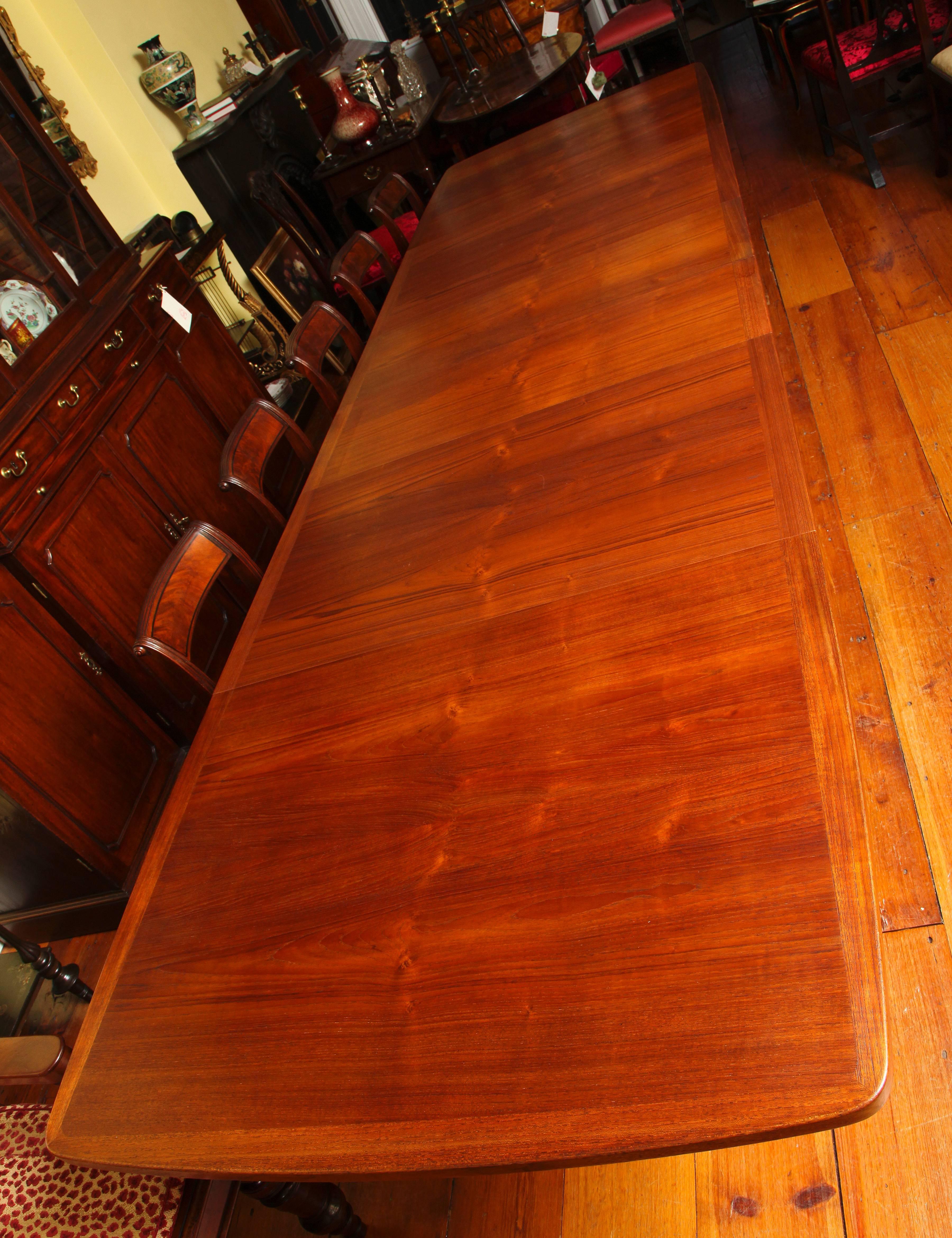 A very elegant and finely figured large teak banquet table, the shaped rectangular top of solid teak with teak banding above a narrow apron and four inset turned tapering legs. The table has four additional original leaves, two 14.5