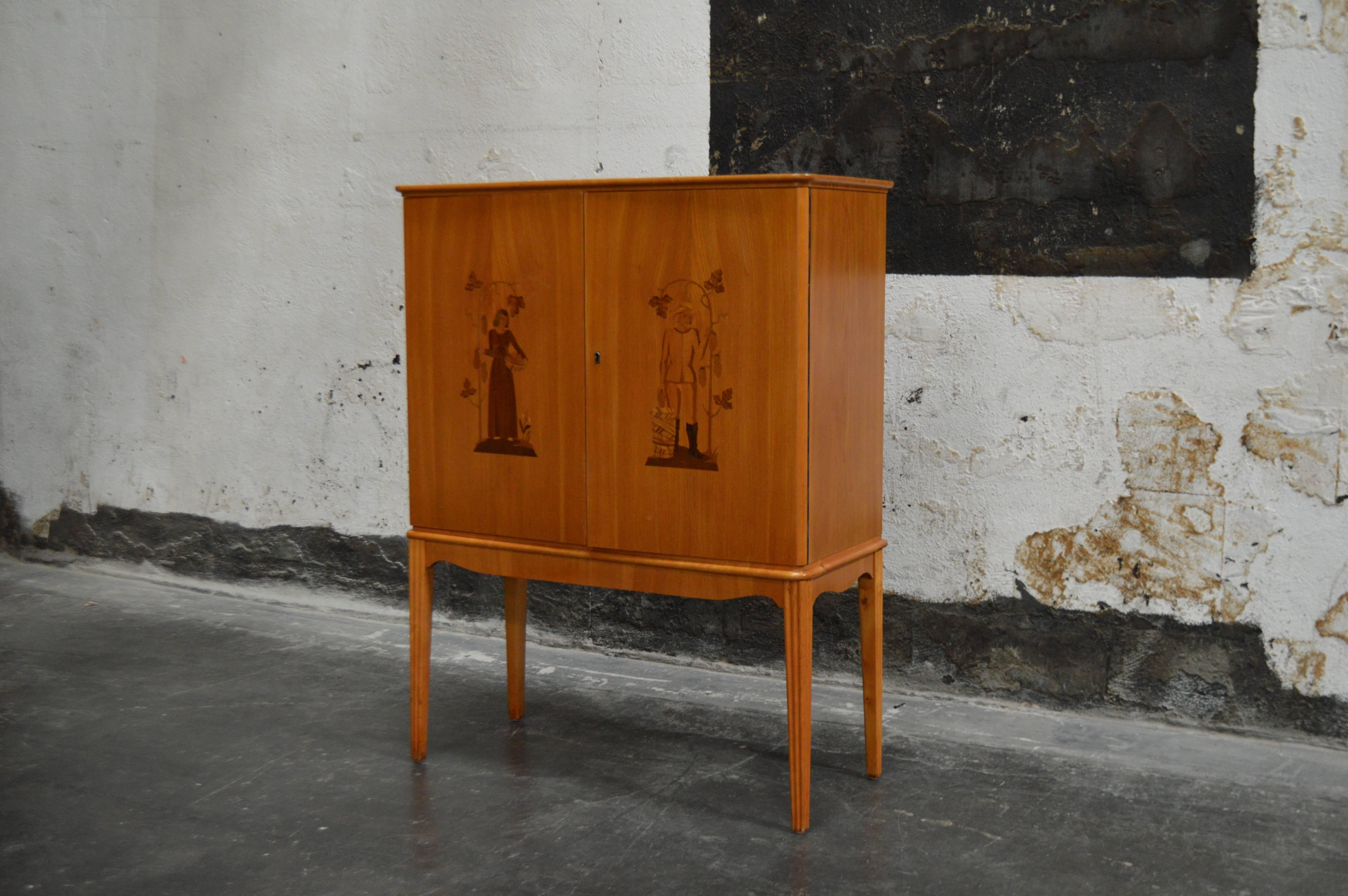 Swedish Art Deco, verging on Art Moderne, storage, bar cabinet in elm, Carpathian elm and dark flame birch with intarsia doors. Upper storage area has adjustable shelving and two interior drawers. Inlaid figures on each upper door. Key included.