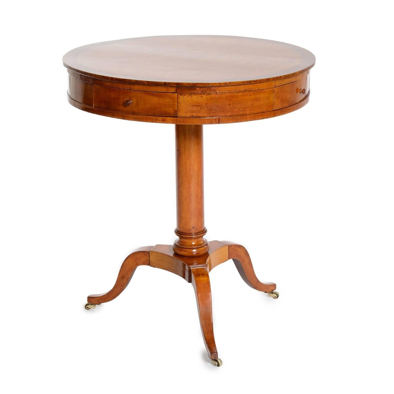 A Swedish Karl Johan birchwood drum table, circa 1830s, the circular top with a birch root crossbanding above a conforming frieze with four drawers, raised on a circular stem with three downswept legs ending with brass-casters.

  