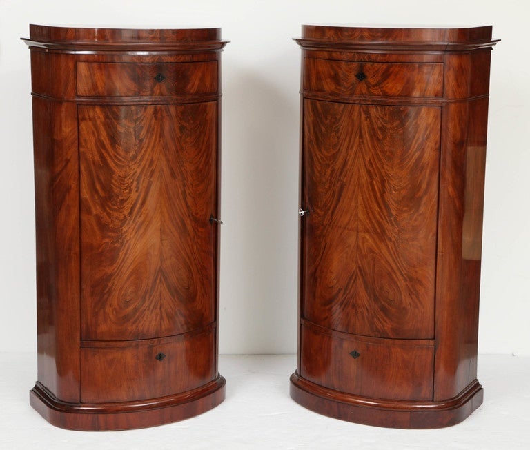 A pair of Danish Empire, from Copenhagen, figured crotch mahogany oval pedestal cabinets, circa 1800-1820, each with an oval stepped top above a molded frieze and a single drawer, the bow-fronted cupboard door and a lower drawer raised on a molded