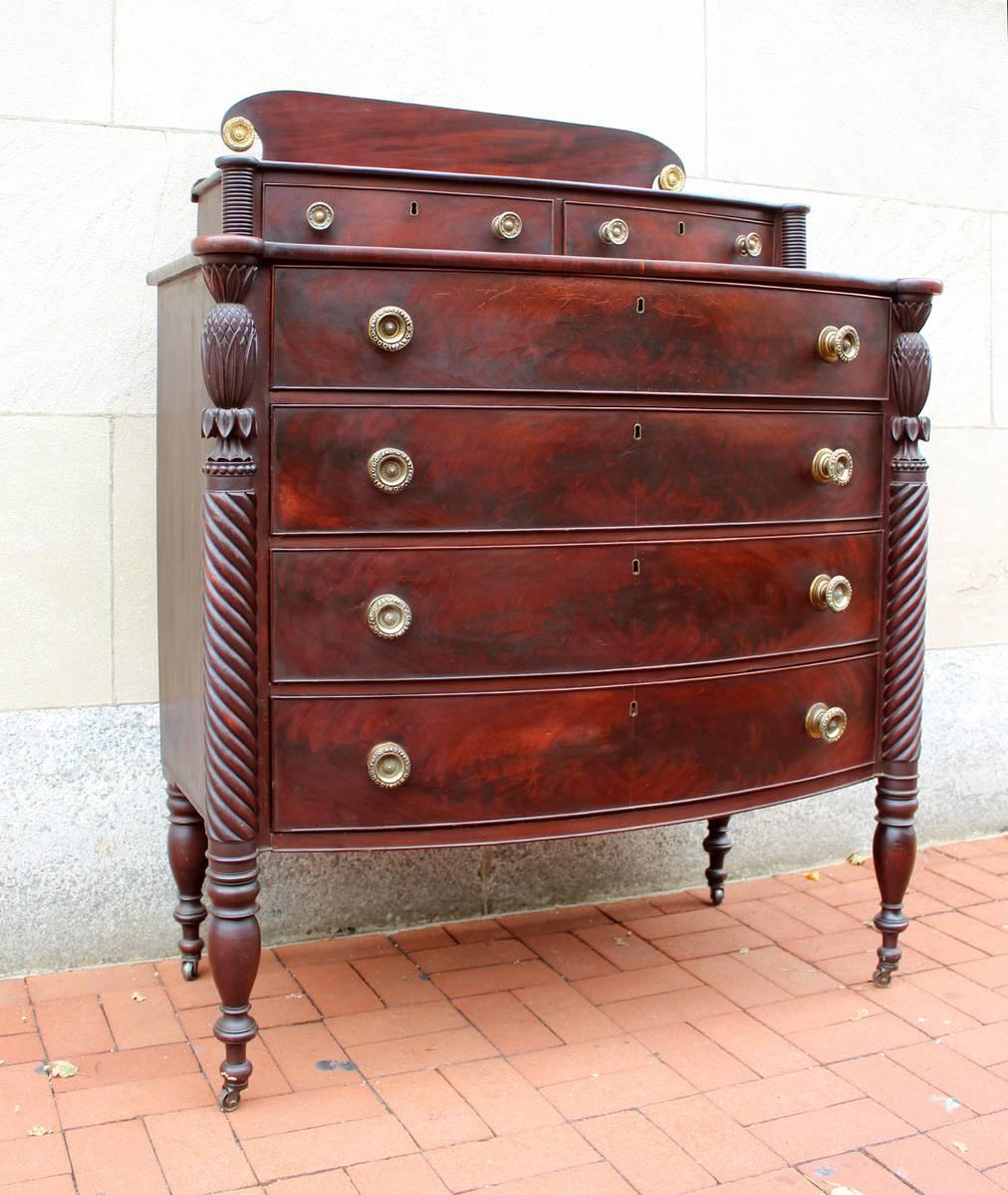 Handsome sheraton mahogany bow-front chest with back splash, excellent grain, fine mirror-image spiraling turned and carved legs with large pineapples and other detailing and original pressed brass rosettes, knobs and castors. North Shore,