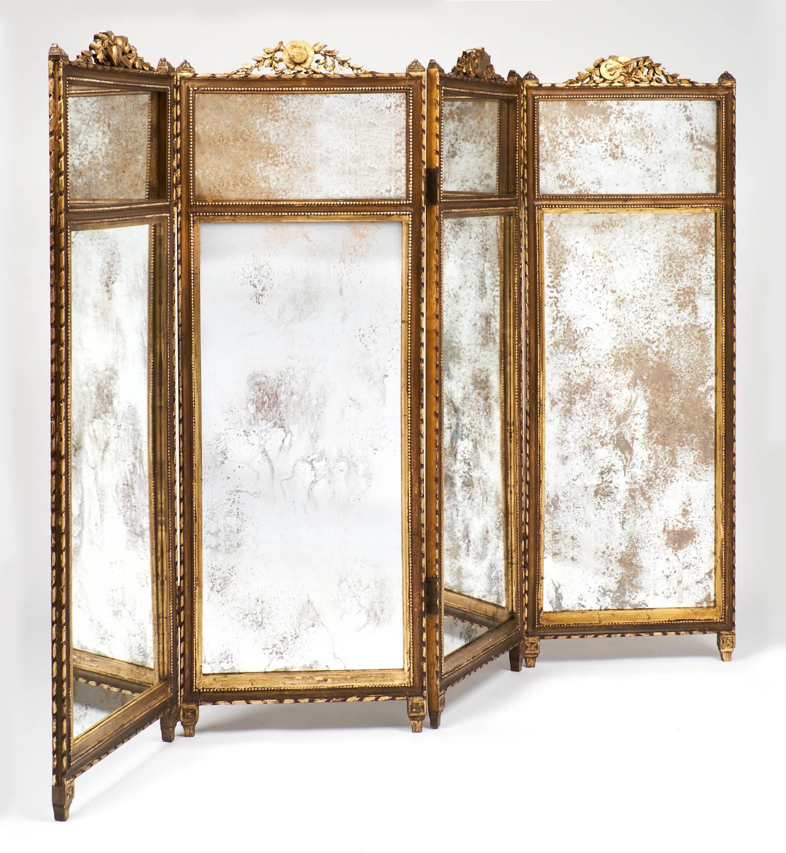 Antique French Louis XVI style folding screen or room divider in four panels of gilded fir with antiqued mirrored glass. Exquisite detailing includes beading around each mirror, and frontons of artistic endeavours surrounded by leaves.