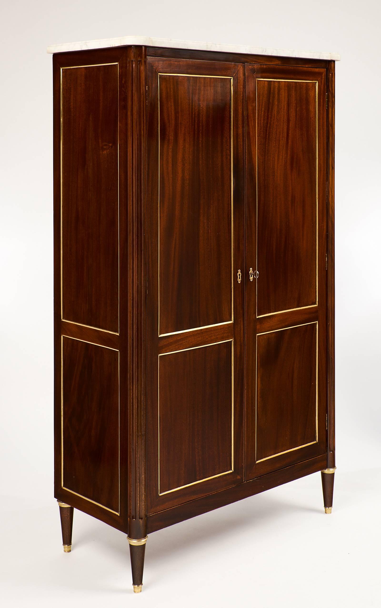 Of flamed mahogany and finished with a lustrous French polish, this stunning armoire features a Carrara marble top, original brass hardware and gilt trims, tapered legs and fluted columns on either side of the doors. The interior shelving makes this