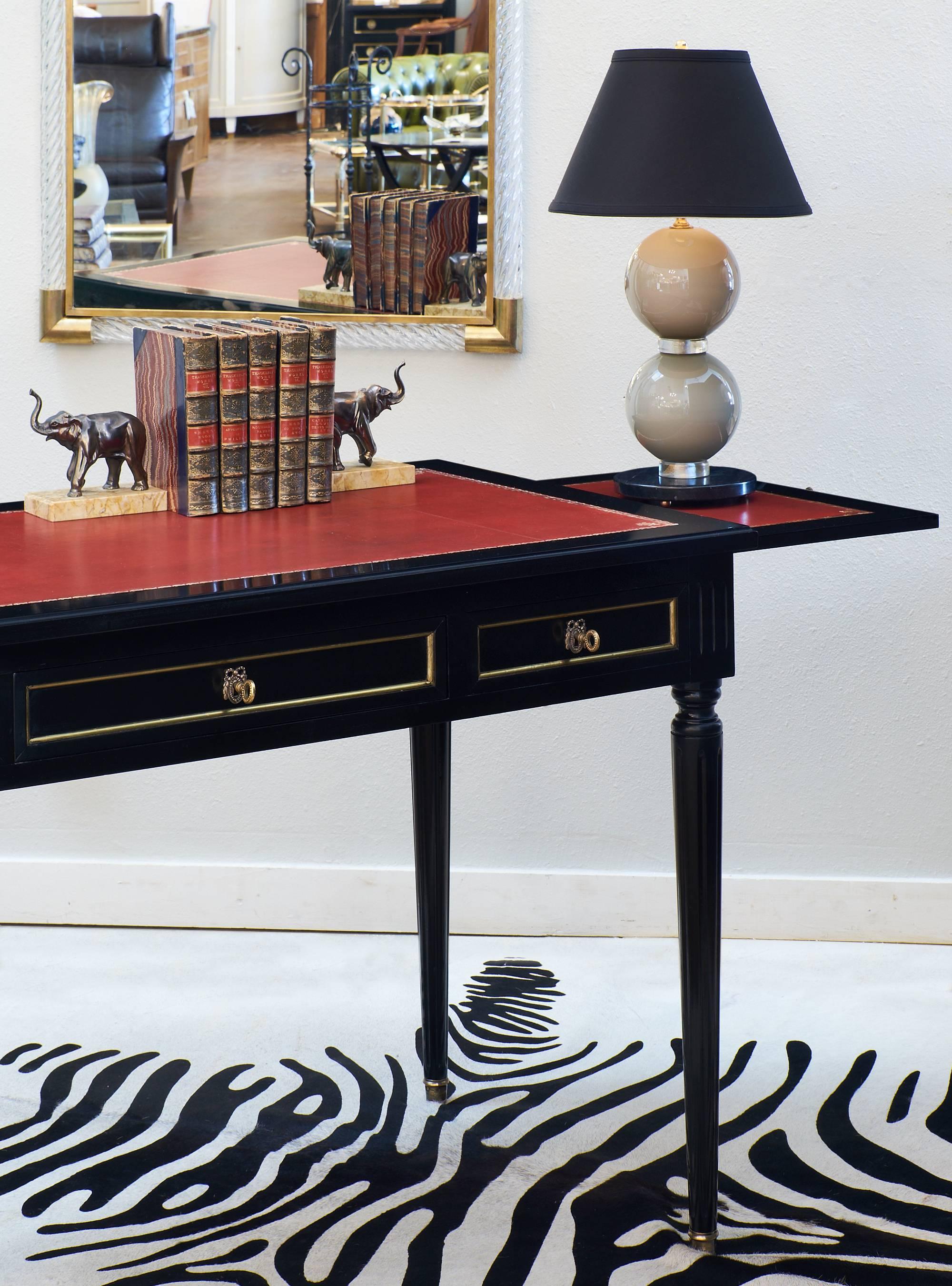 Bureau plat desk in the French Louis XVI style, circa 1940. This elegant writing table has fluted legs, bronze hardware and gilt trim, and three dovetailed drawers. The writing surface is embossed crimson red leather, and covers the two pull-out