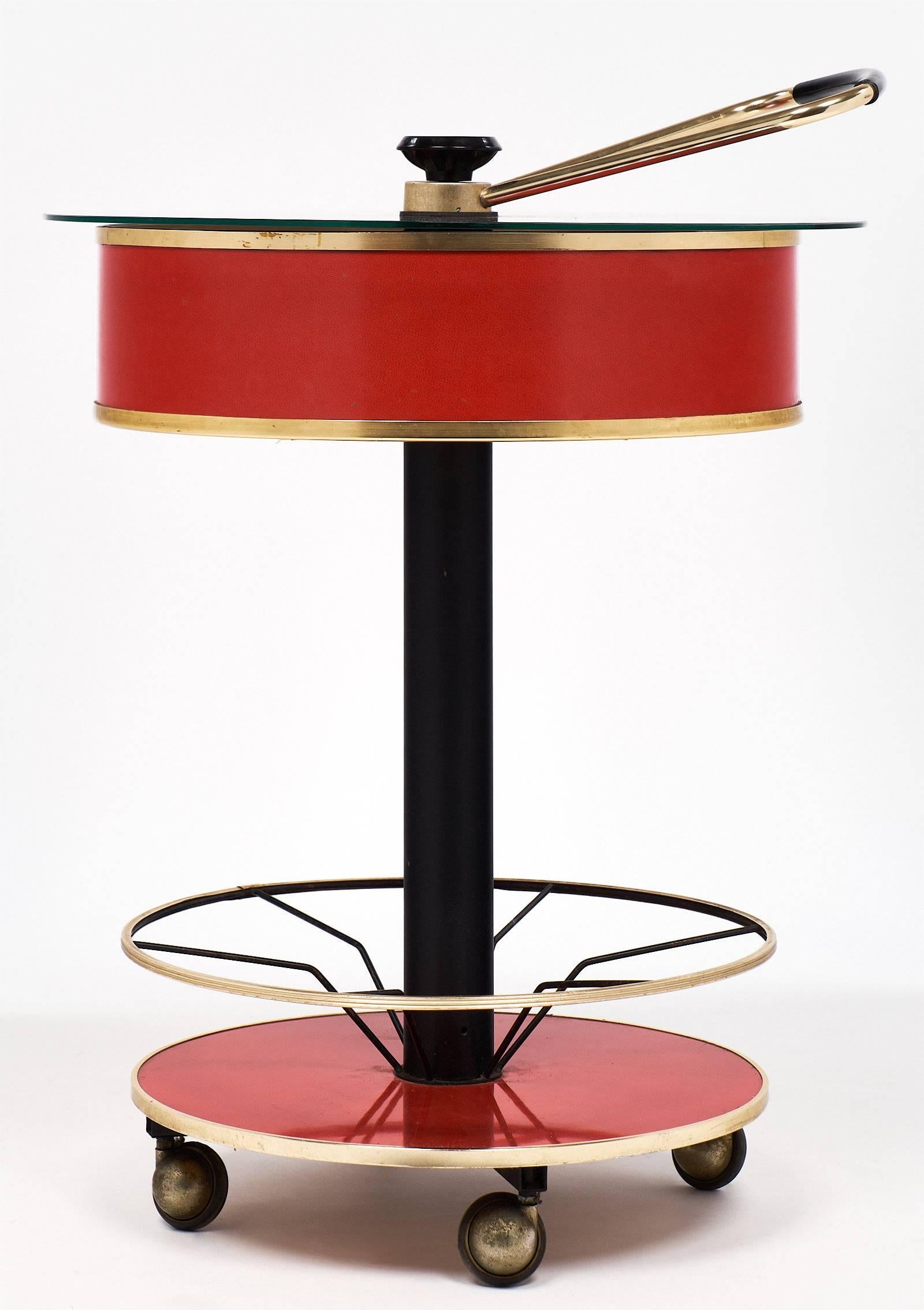 A circular bar cart with whimsical features such as a central leg and a hydraulic system for opening the case using the brass handle. At the raised position, the glass top lifts allowing access for glasses to be stored in the space within. Bottles