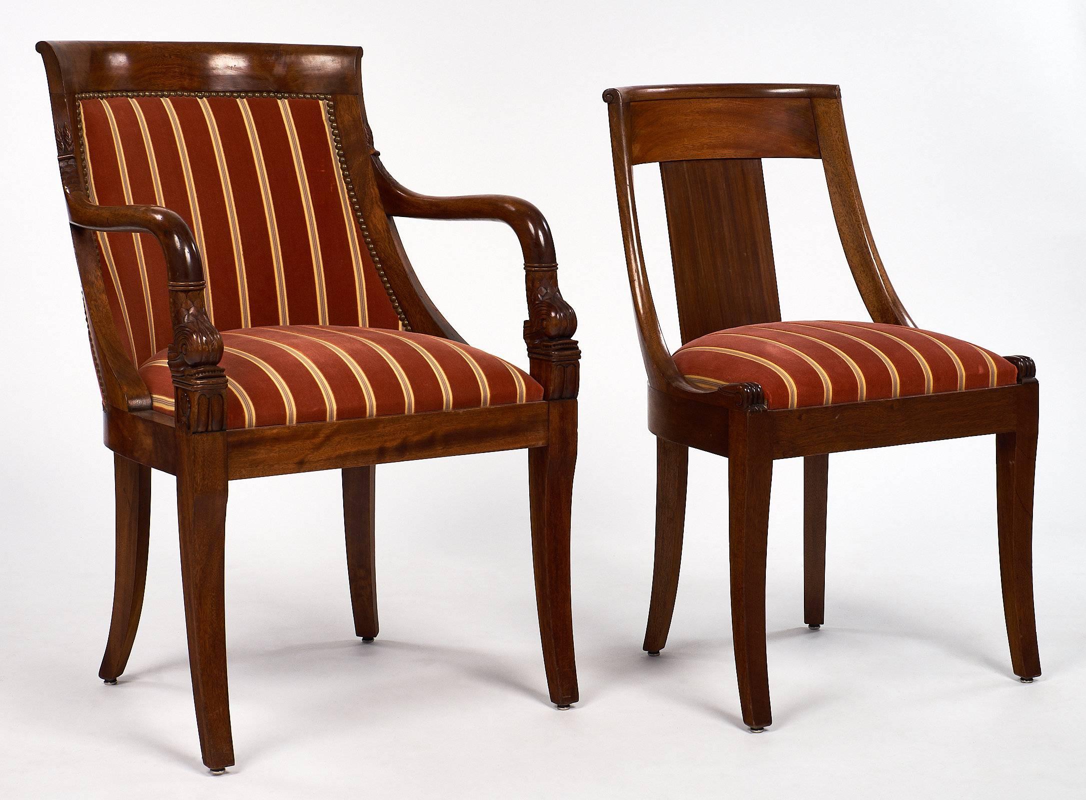 Set of seven Empire style dining chairs. Two of the chairs are armchairs with brass nailheads and beautifully carved mahogany frames. The arms have fantastic detail of stylized fish and scales, and the chairs are barrel shaped. The other five side