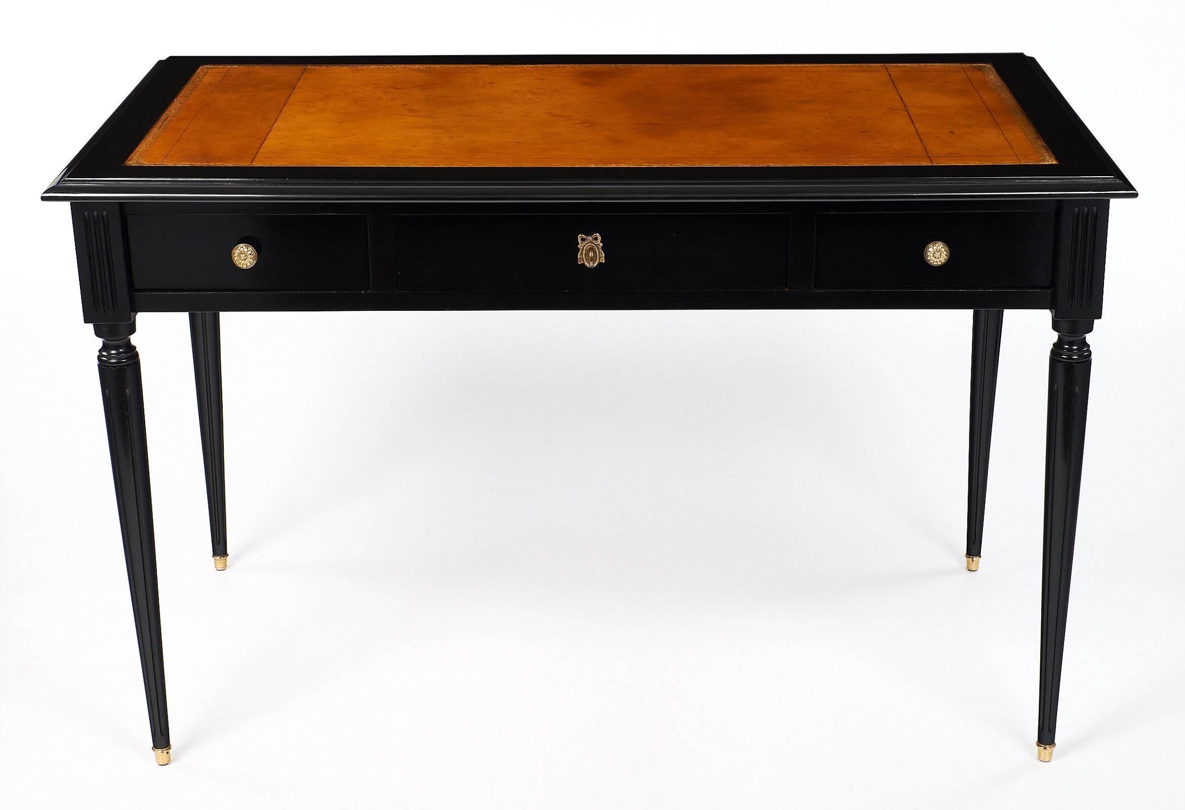 Beautiful writing desk made of ebonized mahogany and finished with a French polish. This great piece has a tan leather writing top, featuring an embossed, gold leaf frieze design. The desk has three drawers on the front side with bronze hardware.