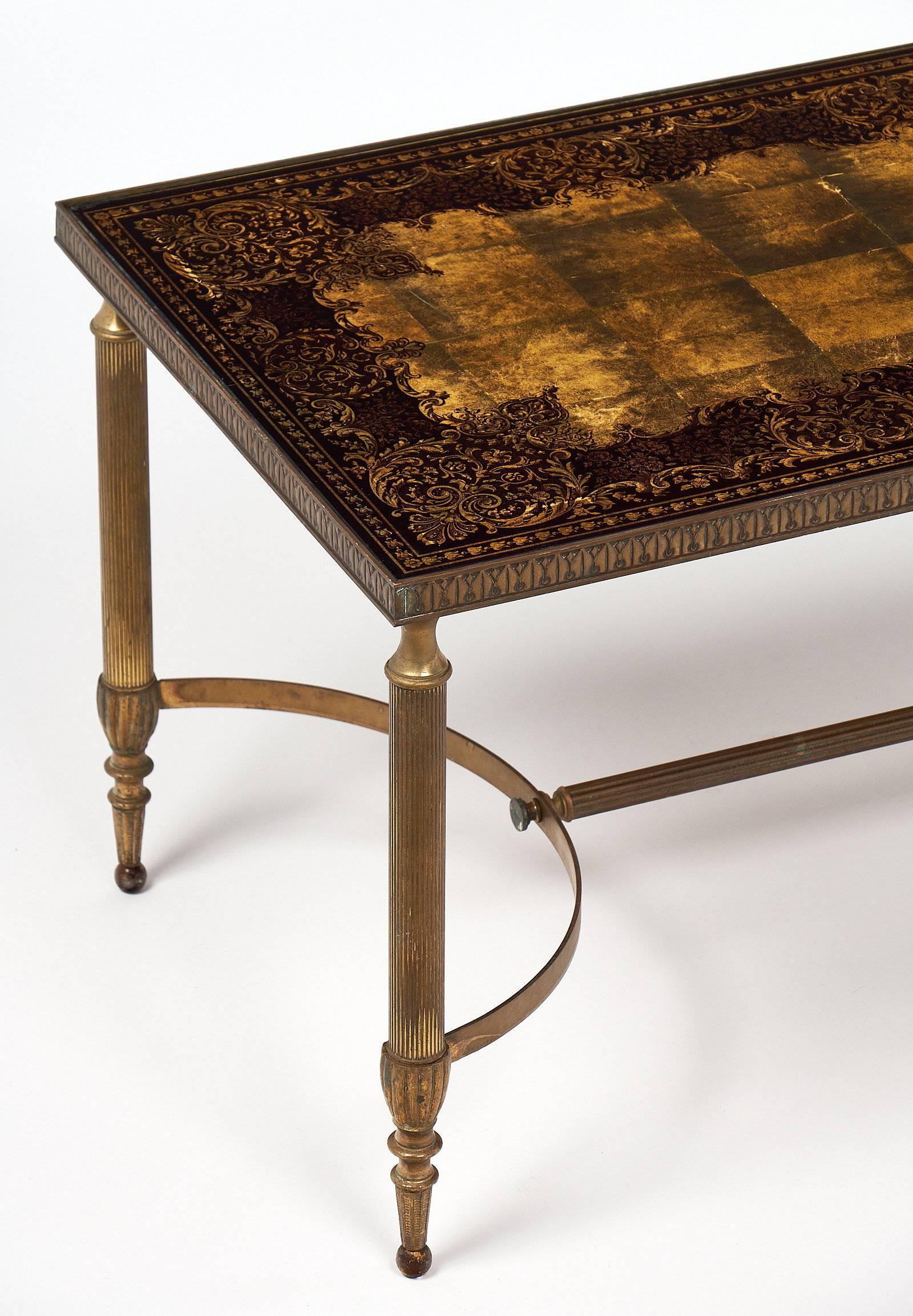 Stunning Art Deco coffee table with a detailed brass base, including stretcher with curved ends. The legs are tapered and fluted. The top is Églomisé glass with a gold and burgundy design including scroll work and floral motifs. This table is an