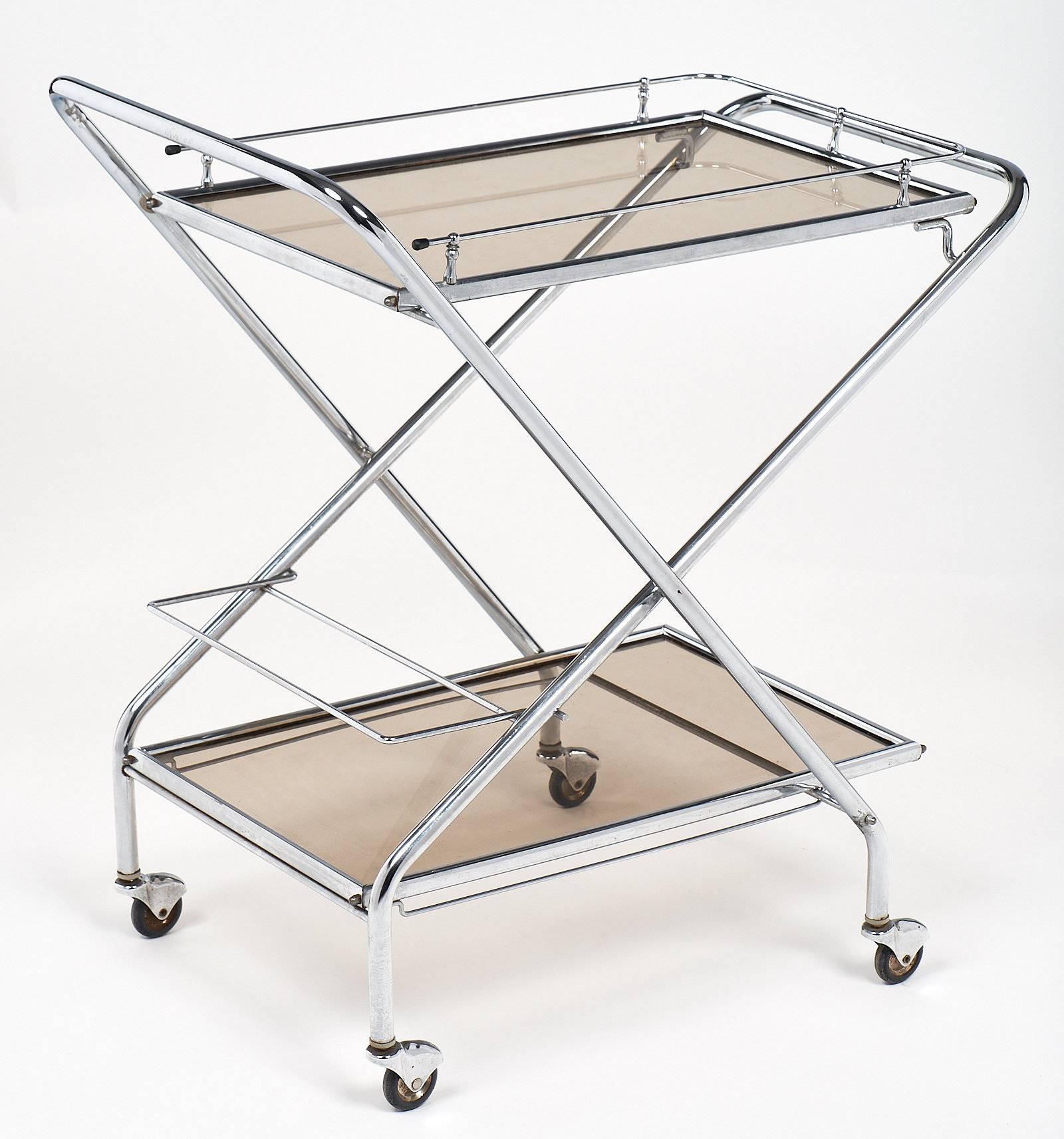 Midcentury folding bar cart on casters in chrome with two smoked glass shelves. The top shelf has a chrome gallery and the bottom shelf contains a single row insert for bottles. A clean and functional piece that will stand the test of time!