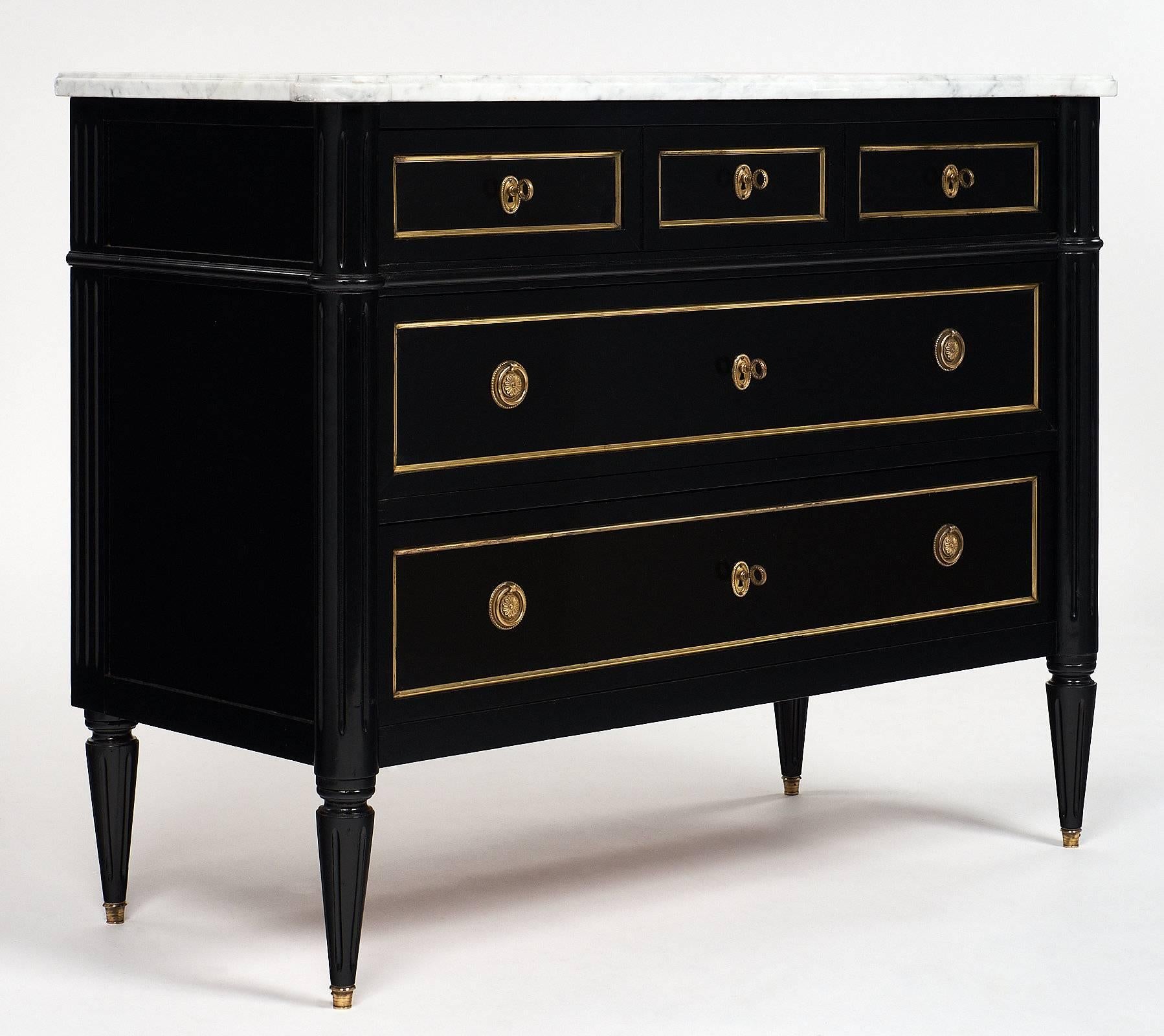 French Louis XVI chest of drawers of mahogany, ebonized and finished with a lustrous French polish, Carrara marble-top, and tapered legs capped with brass feet. The piece has three dovetailed drawers enhanced by gilt brass trims and finely cast