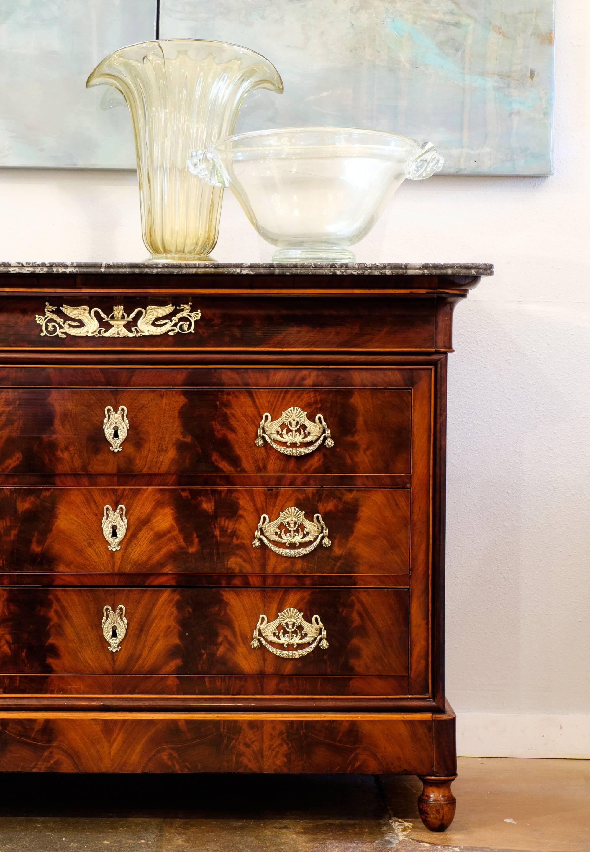 A stunning antique French Restoration period flamed mahogany chest of drawers topped with a beautiful Saint Anne marble top. Detailed with finely cast and pulls, key plates, and locks in bronze doré ormolu. Offers three dovetail drawers for storage.