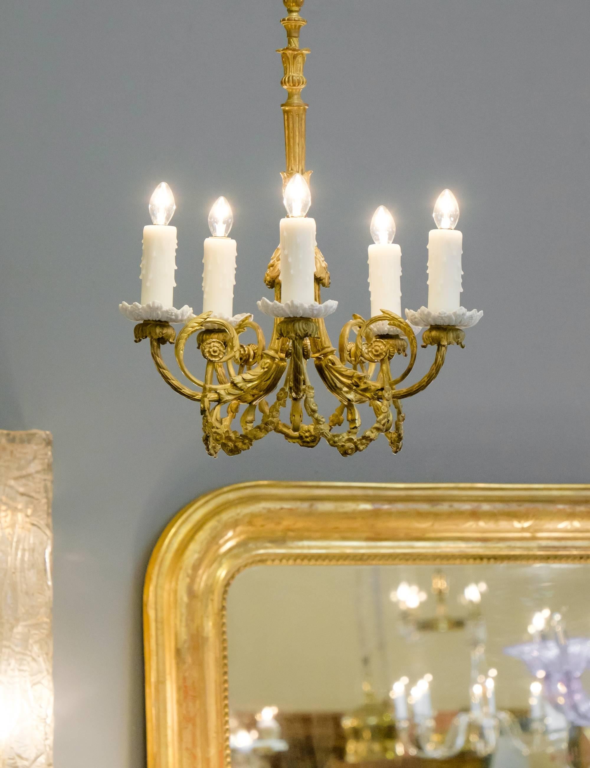 A French 19th century gold-leafed chandelier with porcelain bobeches featuring six arms that have ornately scrolled foliage decoration. This fixture has an equally detailed stem and finial.