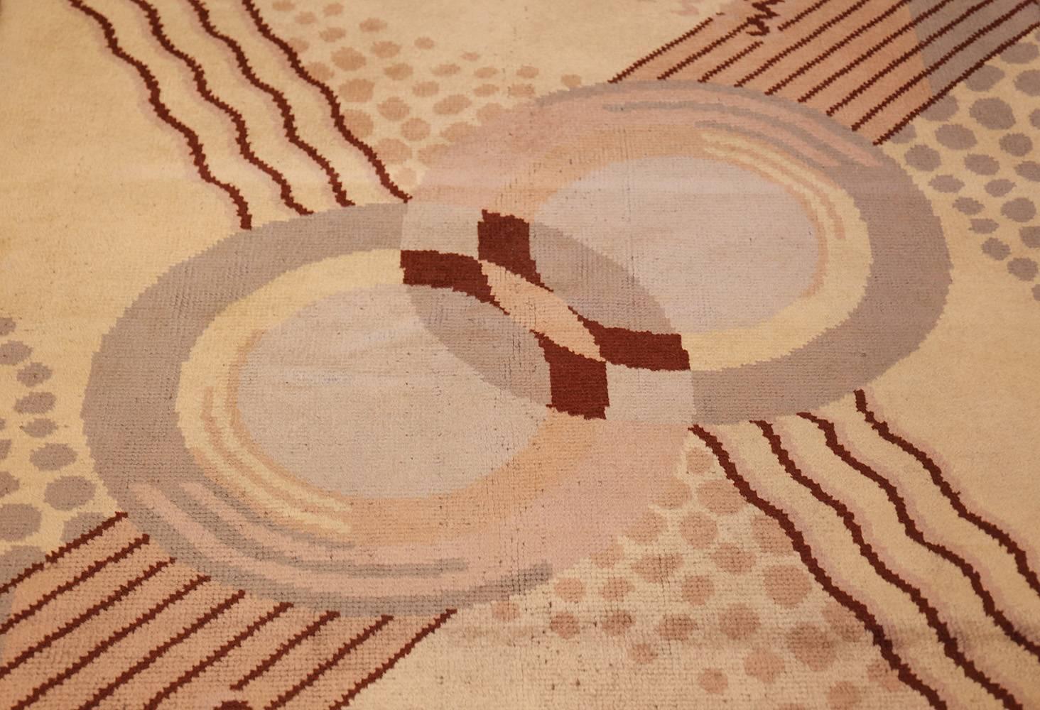 This antique French rug showcases the Art Deco style, featuring a light tan and cream body with red accents. The design centers around a pair of interlocking circles, each fixed with a solid white, round shape in the middle. Where their circular